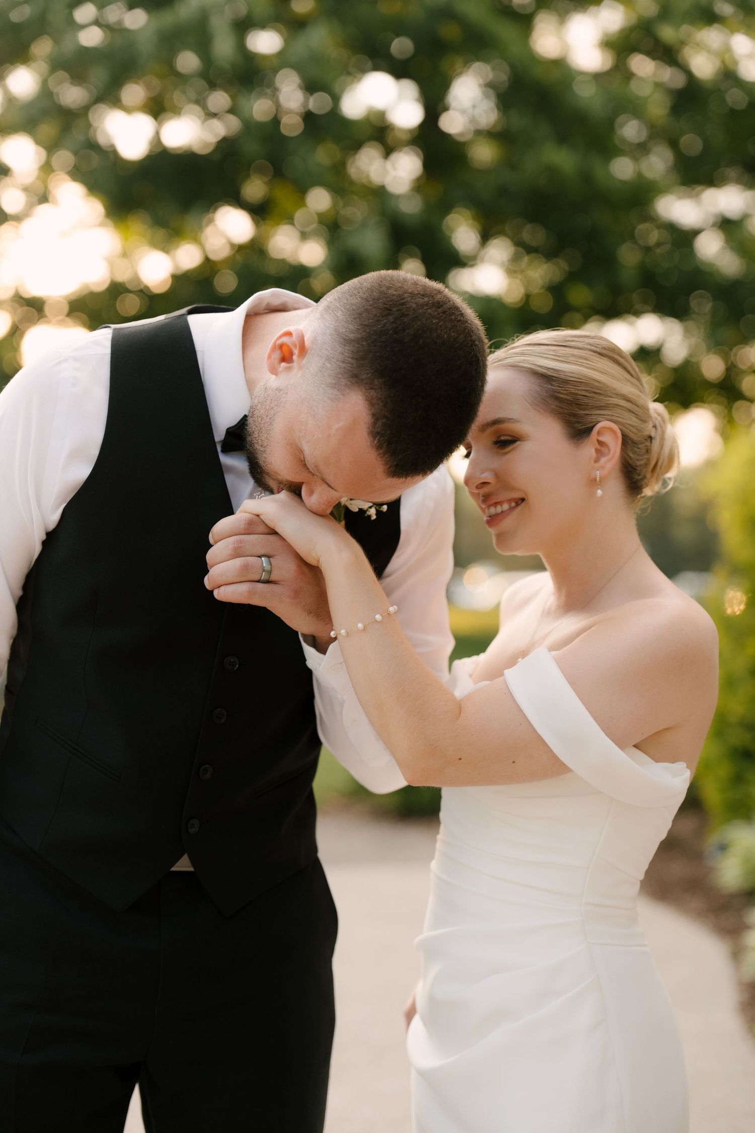 Groom kissing bride's hand tenderly while she smiles, wearing off-shoulder white gown in outdoor garden setting in Guelph.