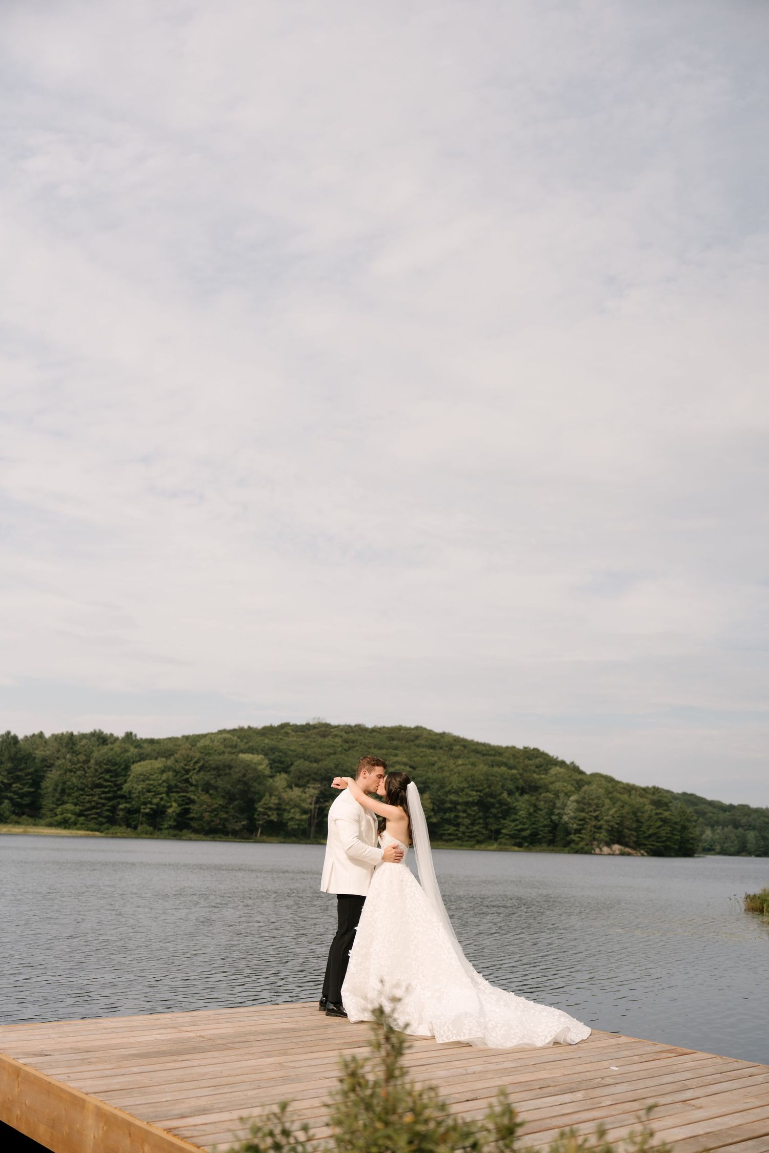 Bride and groom share a romantic kiss on a wooden dock with a serene lake and forested hills in the background.