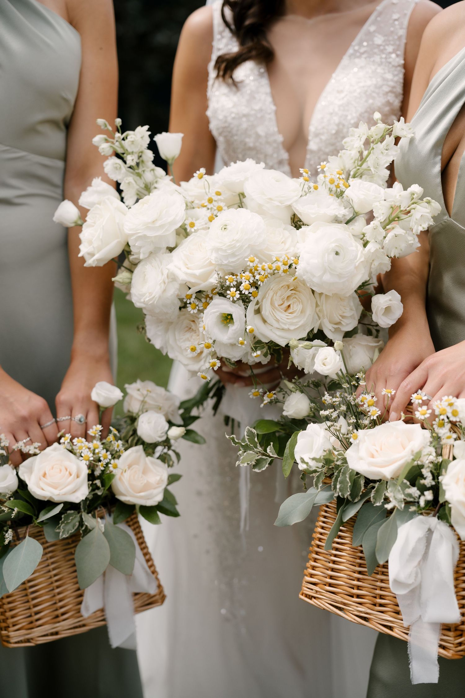 Bride holding large white floral bouquet with bridesmaids carrying wicker baskets filled with white roses and greenery.