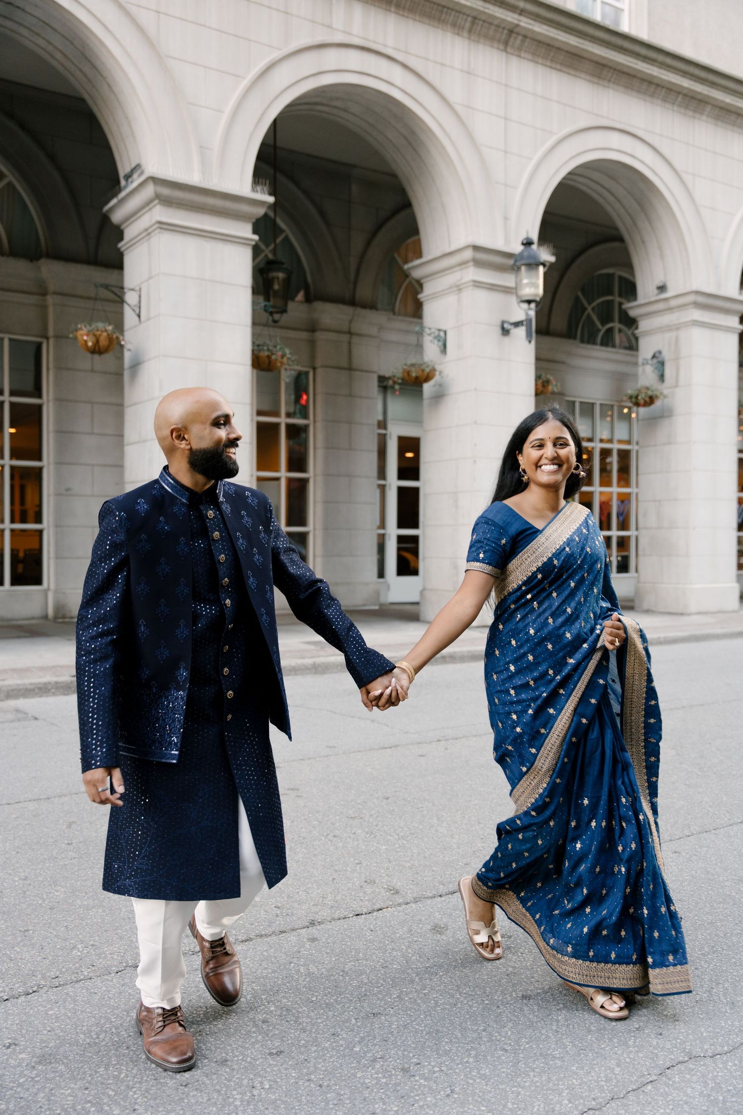 Couple in matching navy blue Indian wedding attire holding hands and walking outside a white stone building.