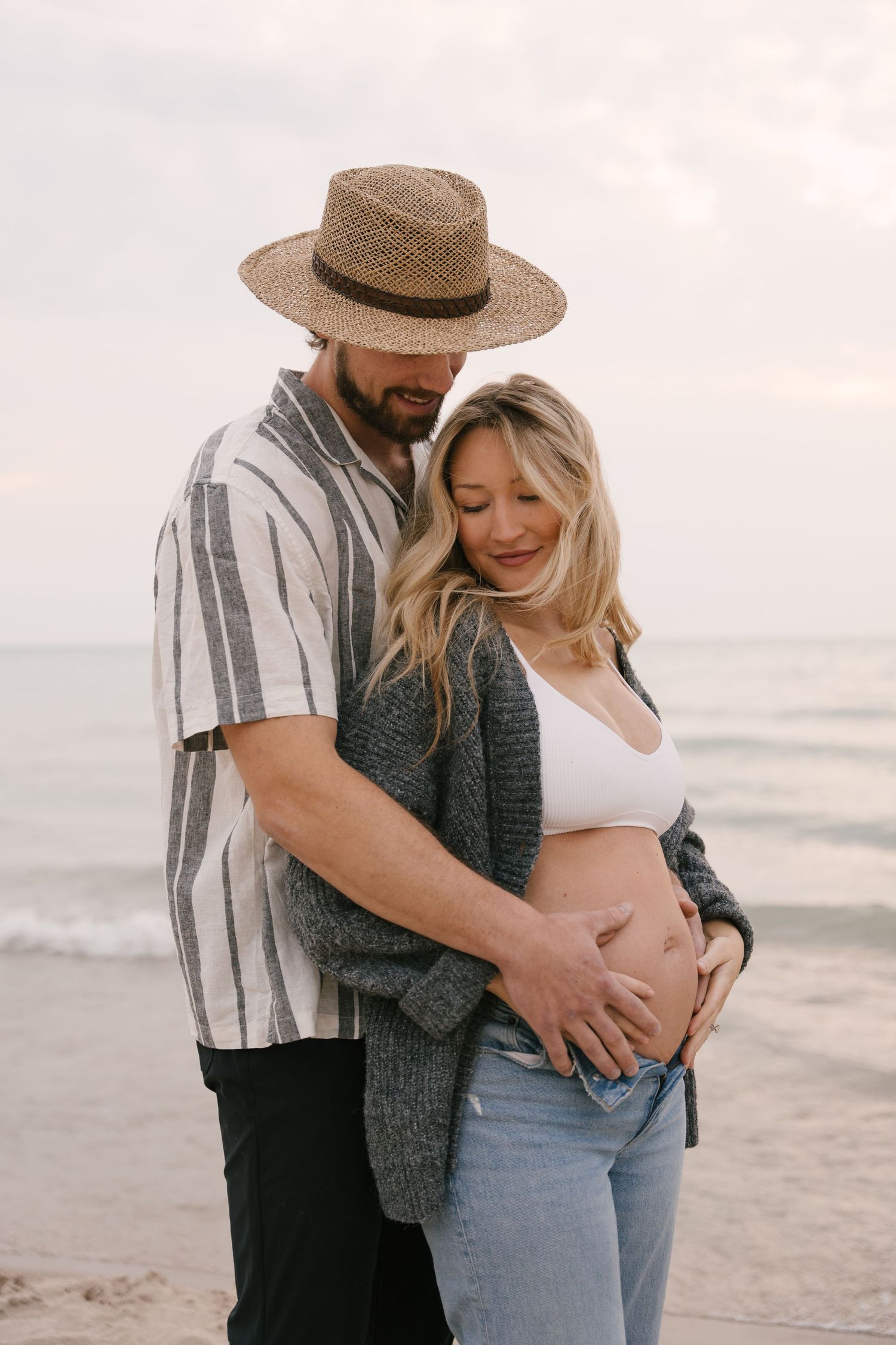 Expecting couple on sandy beach at dusk, man in straw hat hugging pregnant partners belly.