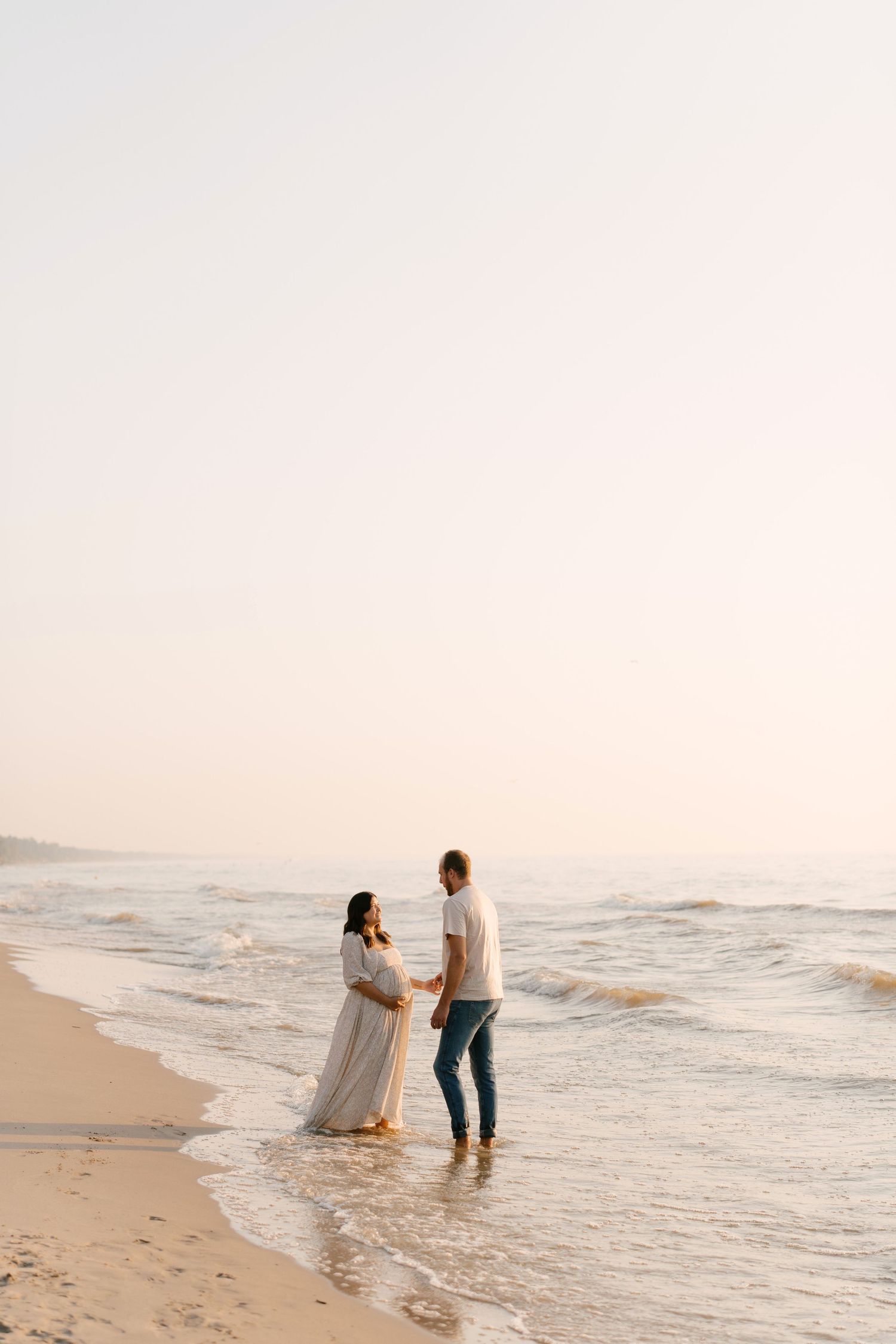 Expectant couple holding hands facing ocean waves at sunset on empty sandy beach.