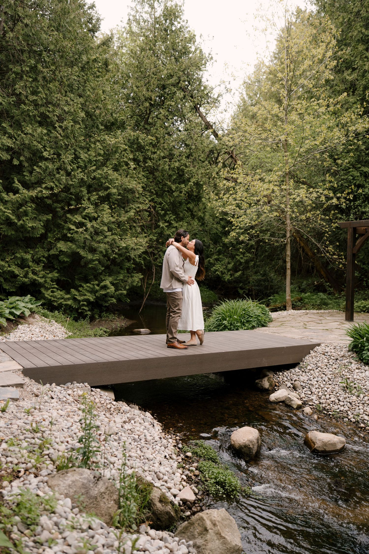Couple embracing and kissing on a wooden bridge over a rocky stream surrounded by lush green trees.