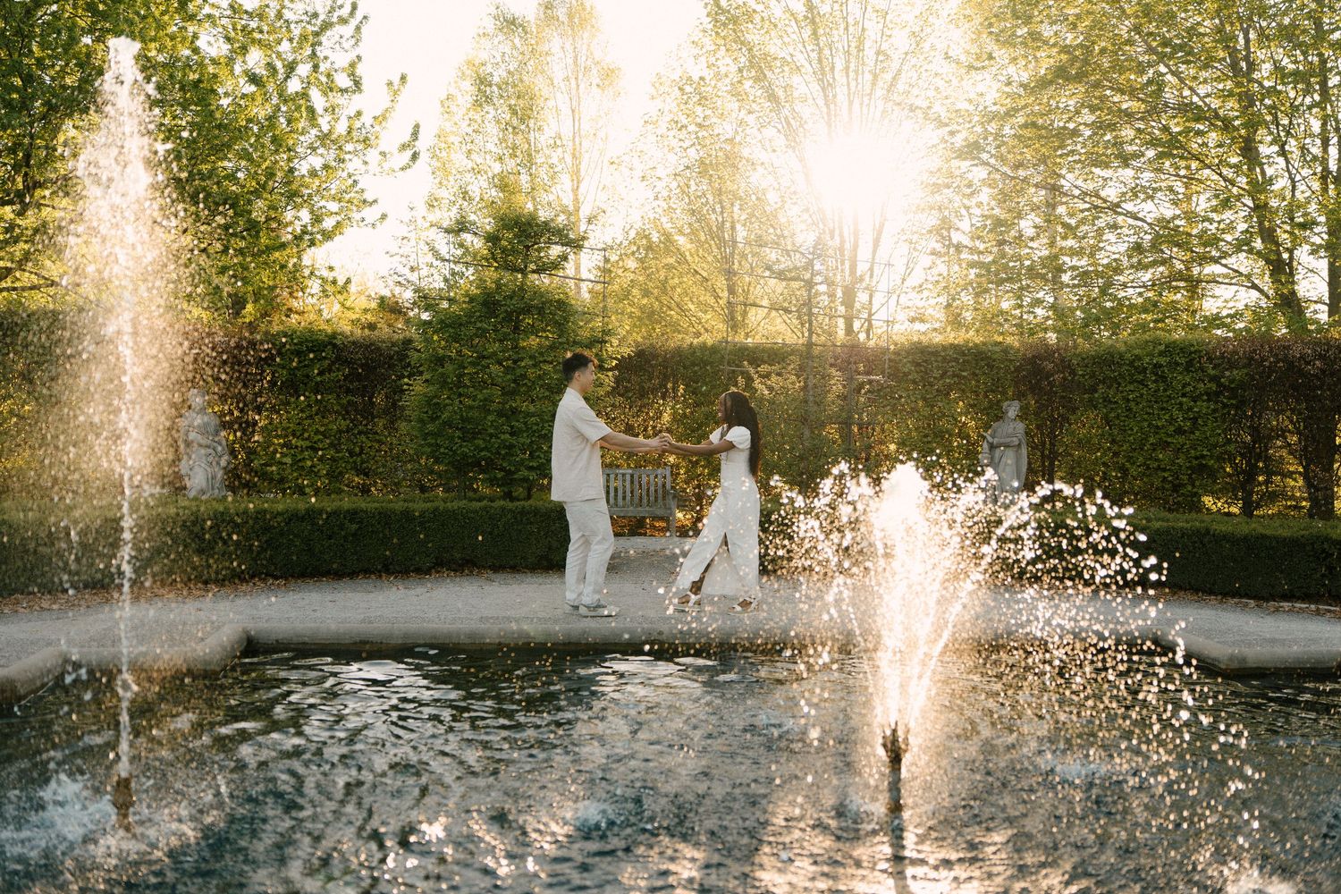Couple holding hands walking past fountain jets in a sunlit formal garden at golden hour.