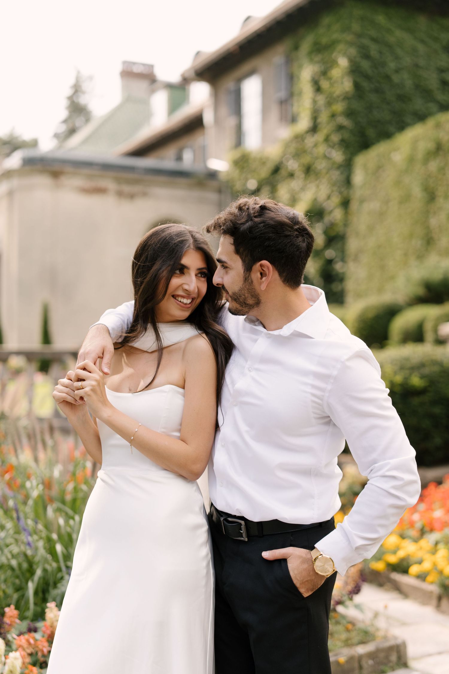 Smiling couple embracing in a garden with ivy-covered estate building in background.
