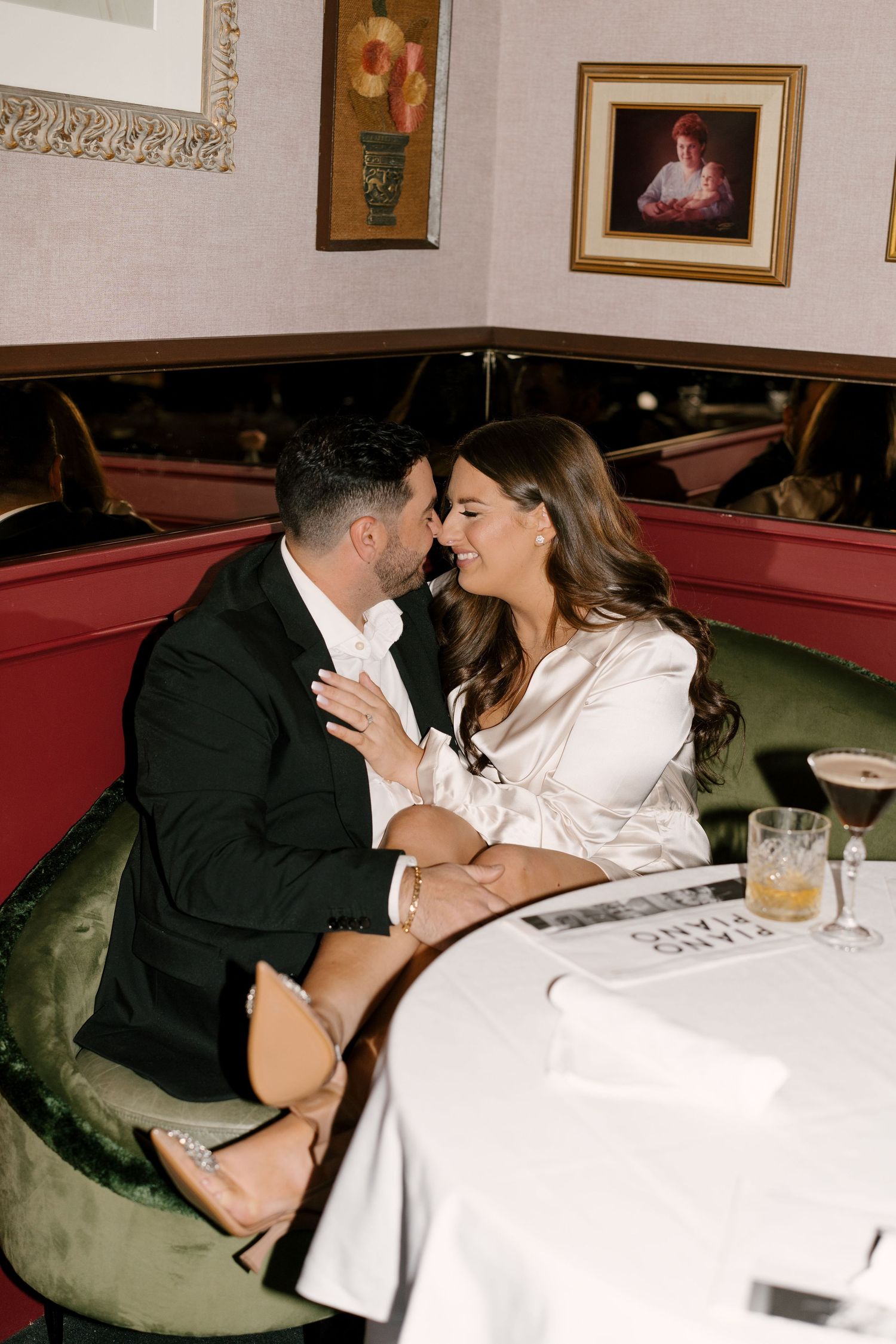 Romantic couple sharing an intimate moment in green velvet booth at upscale restaurant with cocktails on table.