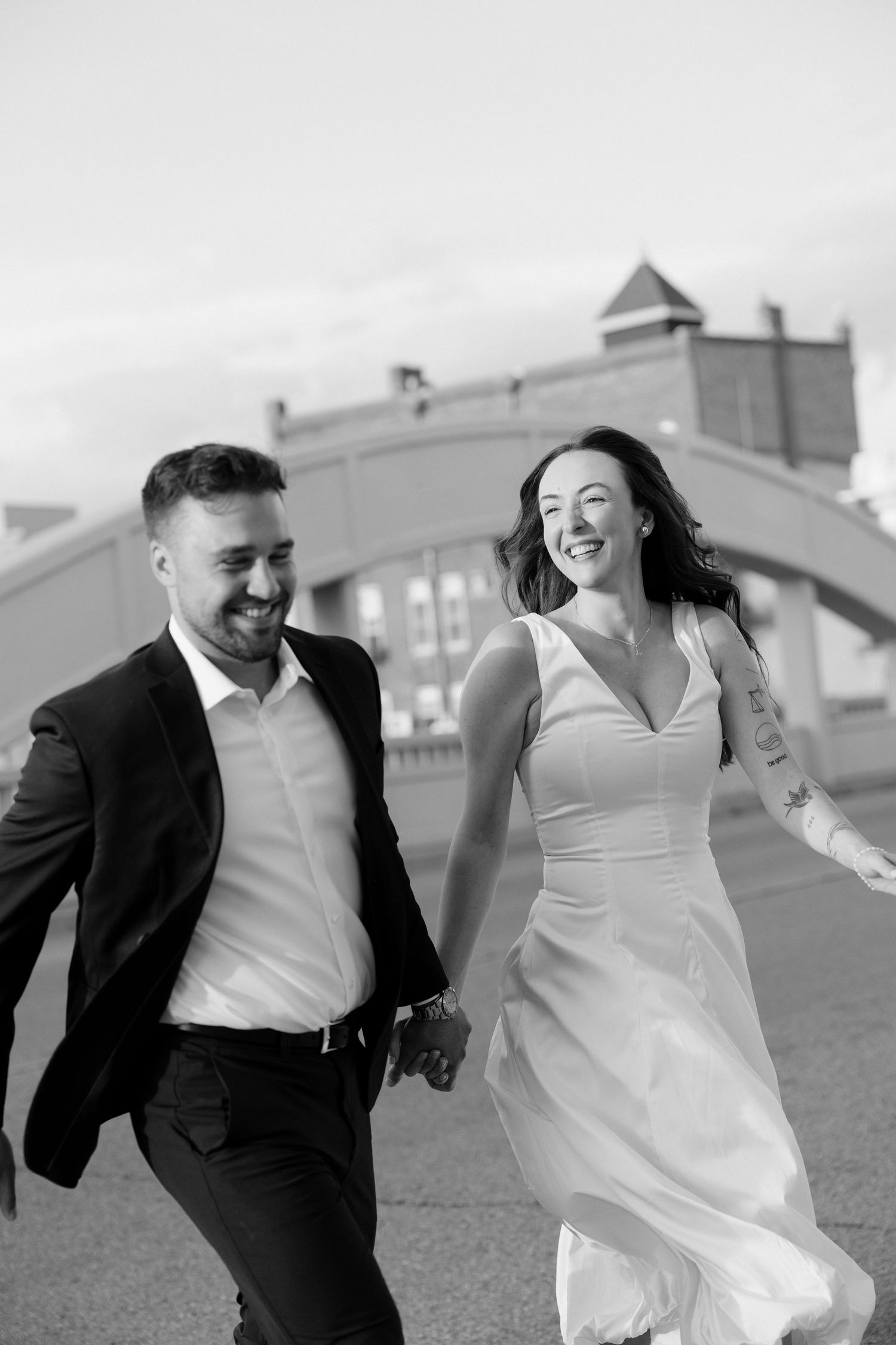 Black and white photo of laughing couple running hand in hand in wedding attire outdoors.