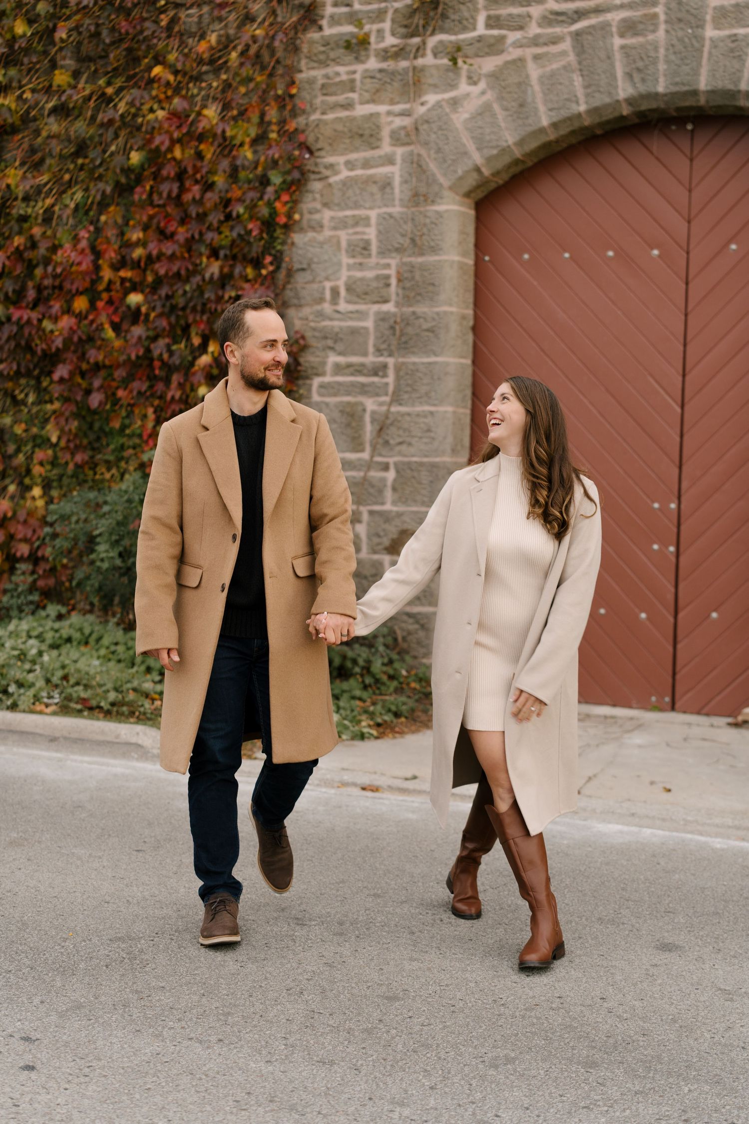 Smiling couple in matching camel coats holding hands and walking toward camera by stone building.