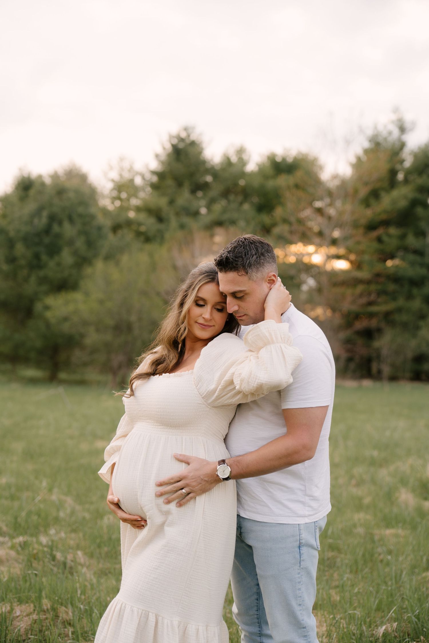 Pregnant couple embracing in a green meadow at golden hour, woman in white off-shoulder dress.