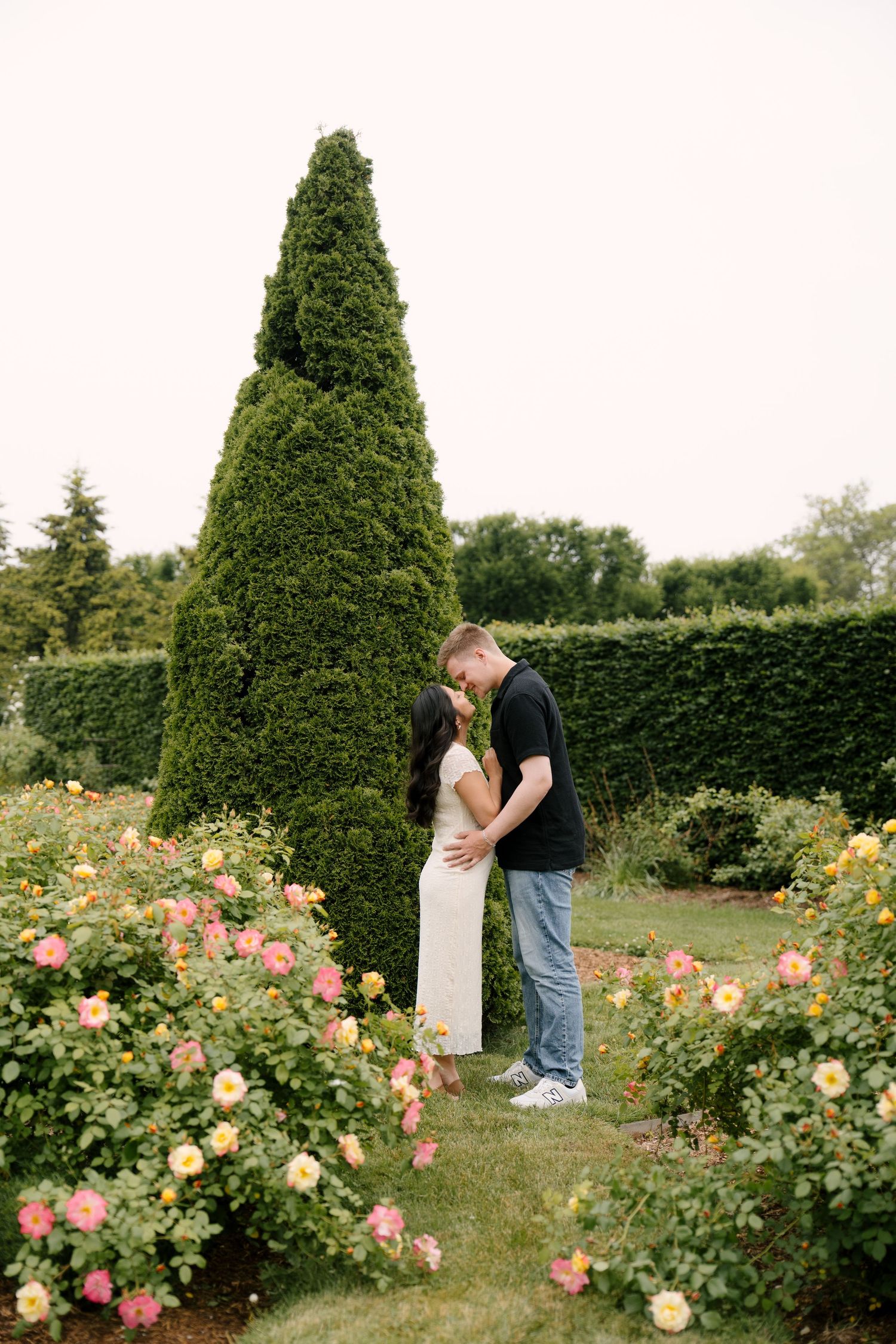 Couple kissing beside a tall evergreen tree surrounded by colorful roses in a lush garden.