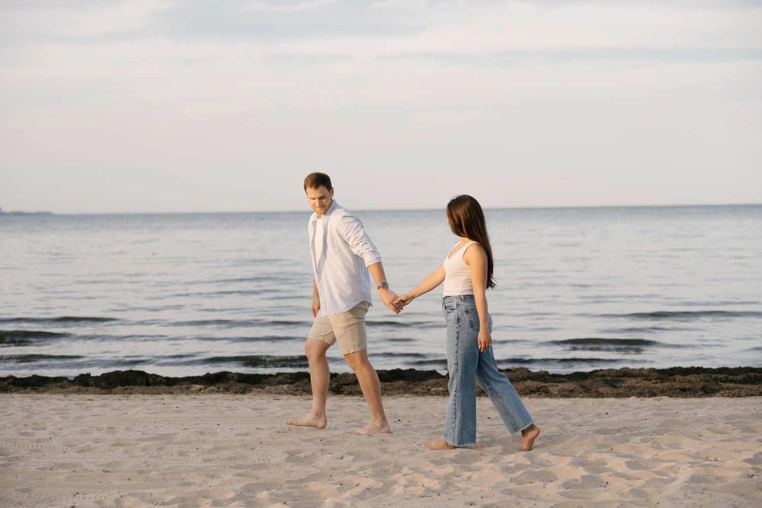 Couple holding hands walking along sandy beach shoreline at dusk, woman in jeans and white top.
