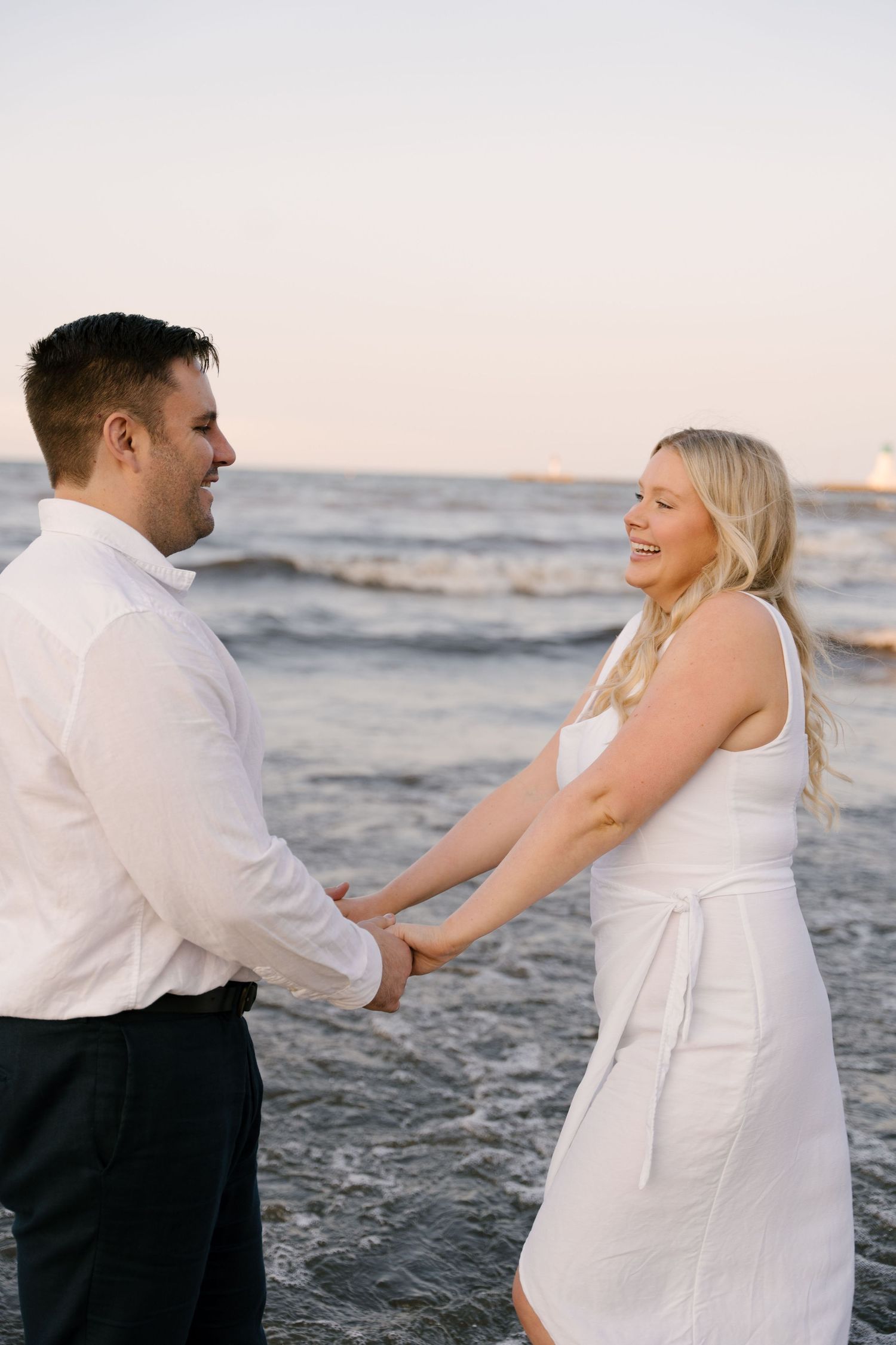Happy couple in white outfits holding hands and laughing together at the ocean's edge at sunset.