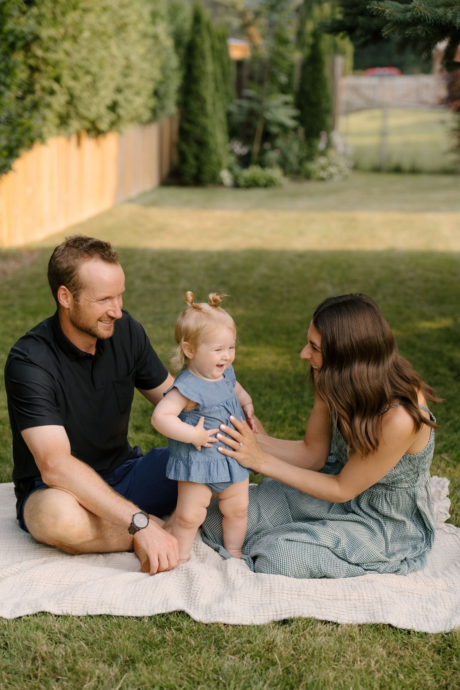 A joyful family sits on a blanket in a sunny backyard with their laughing toddler in a blue dress.