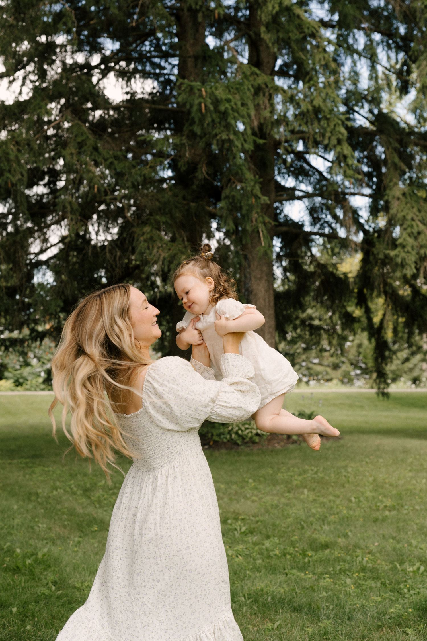 Mother in white dress lifting laughing toddler in sunny green park.