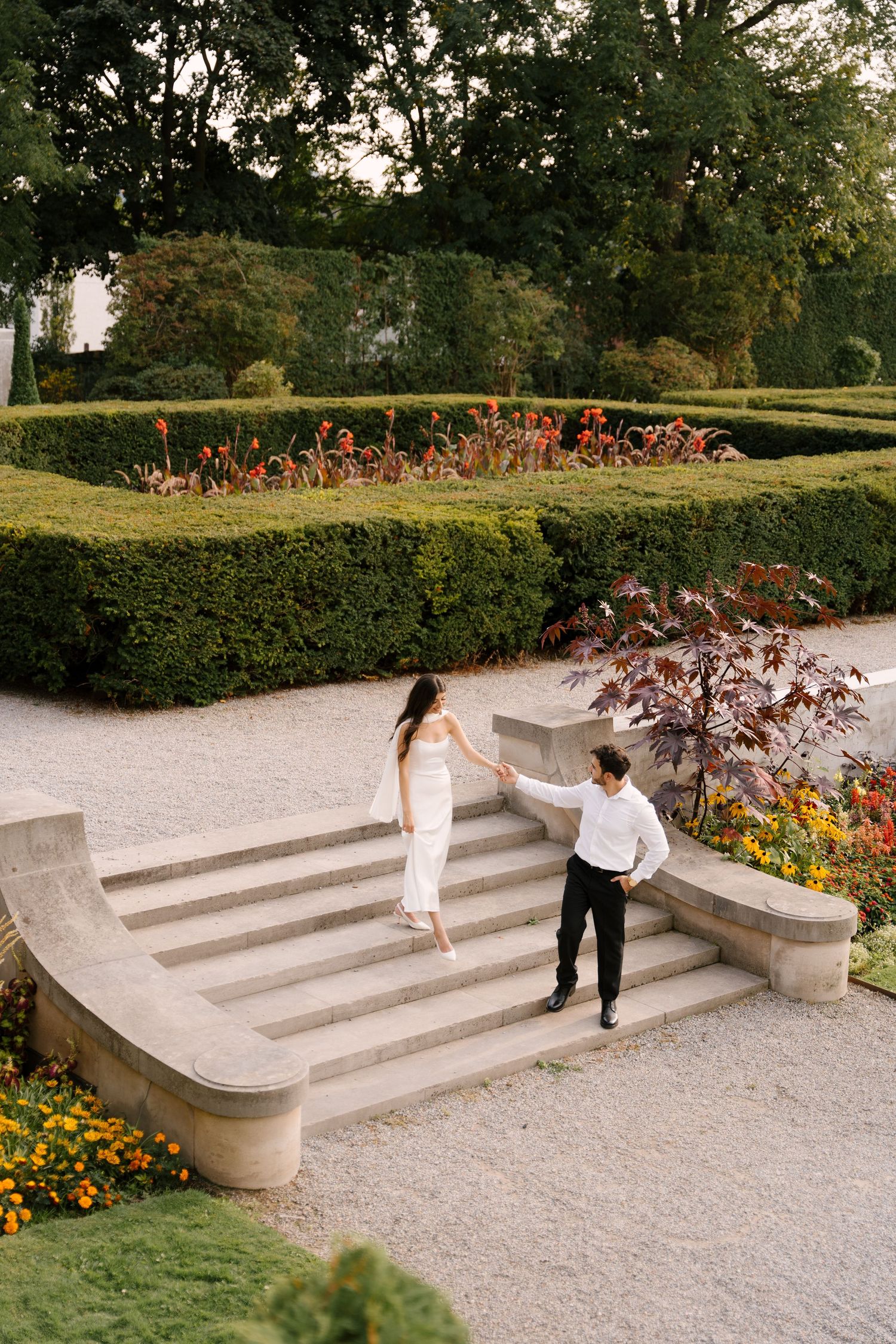 Romantic couple holding hands on curved stone steps surrounded by manicured hedges and colorful flowers.