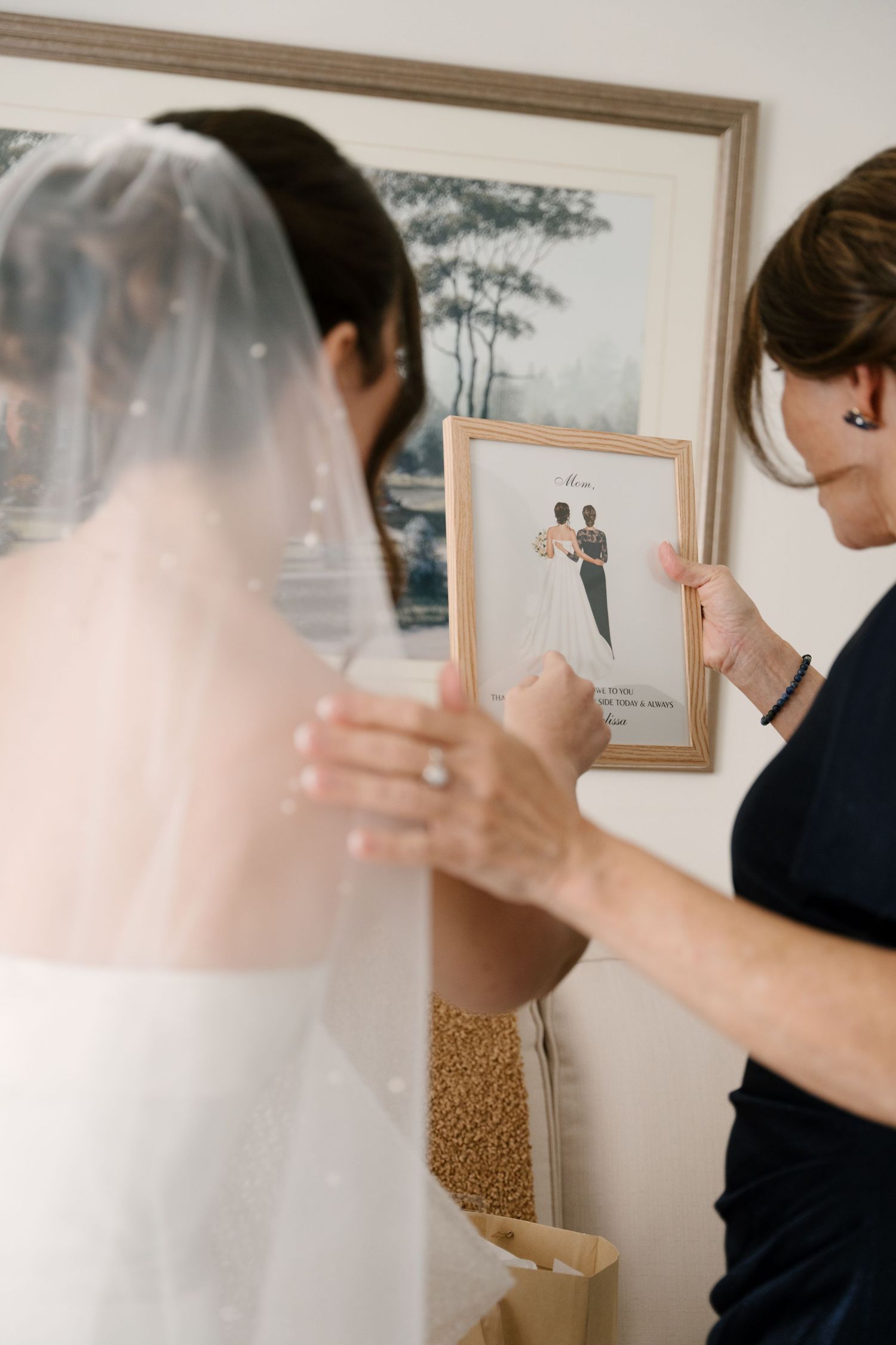 Mother holding a framed illustrated portrait next to a bride in a veil and white wedding gown.