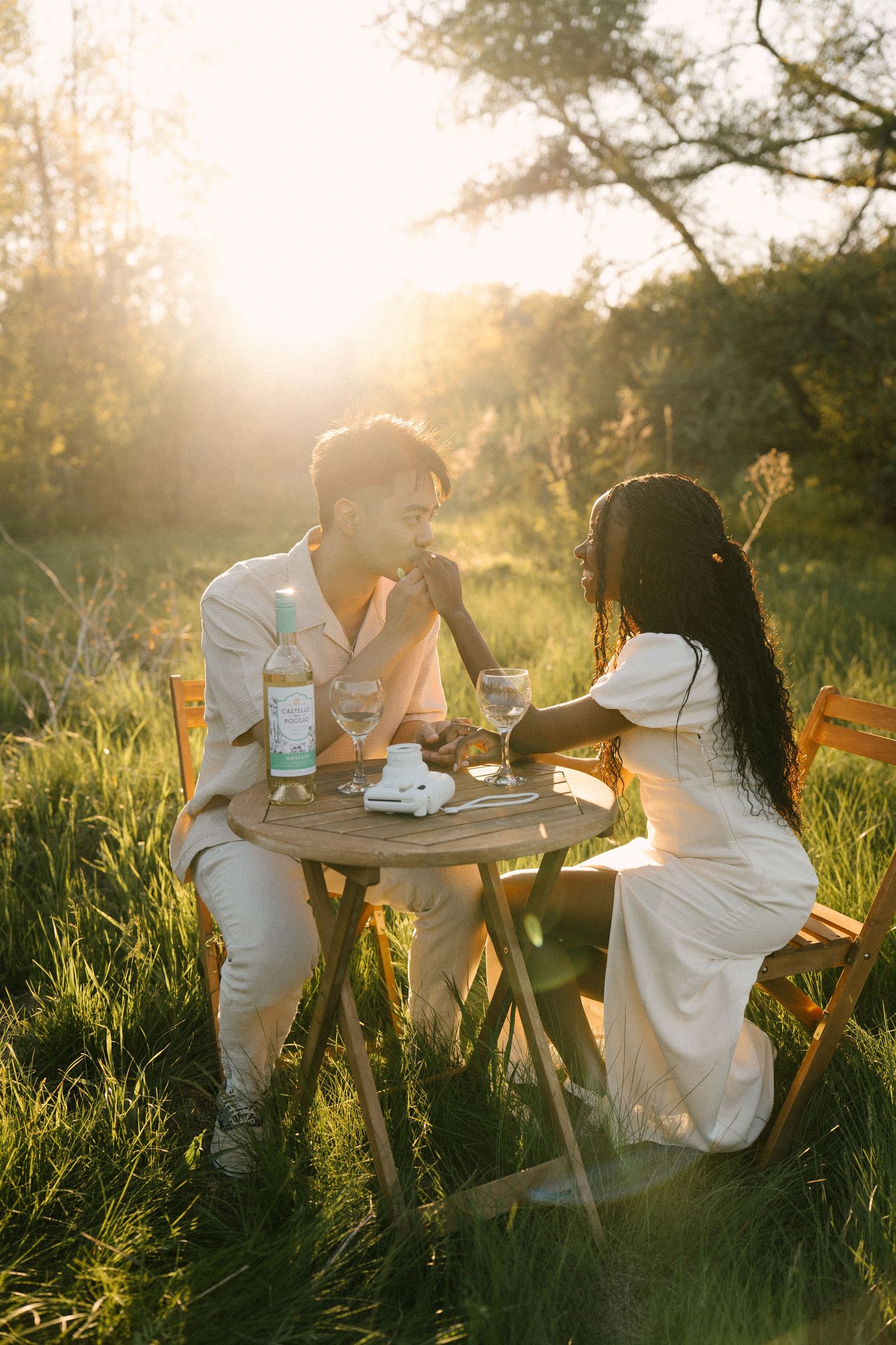 Romantic interracial couple sitting at a wooden table in a sunlit meadow, sharing wine and conversation at golden hour.