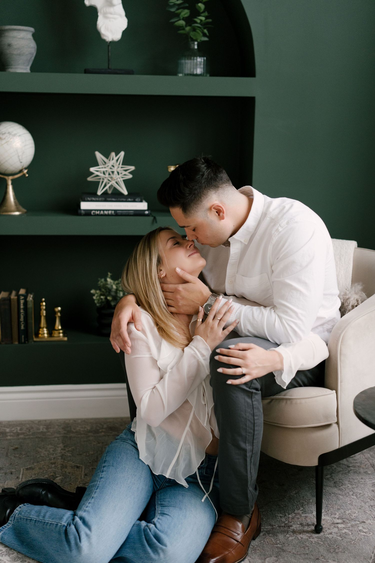 Man kissing blonde woman beside green bookshelf in studio.