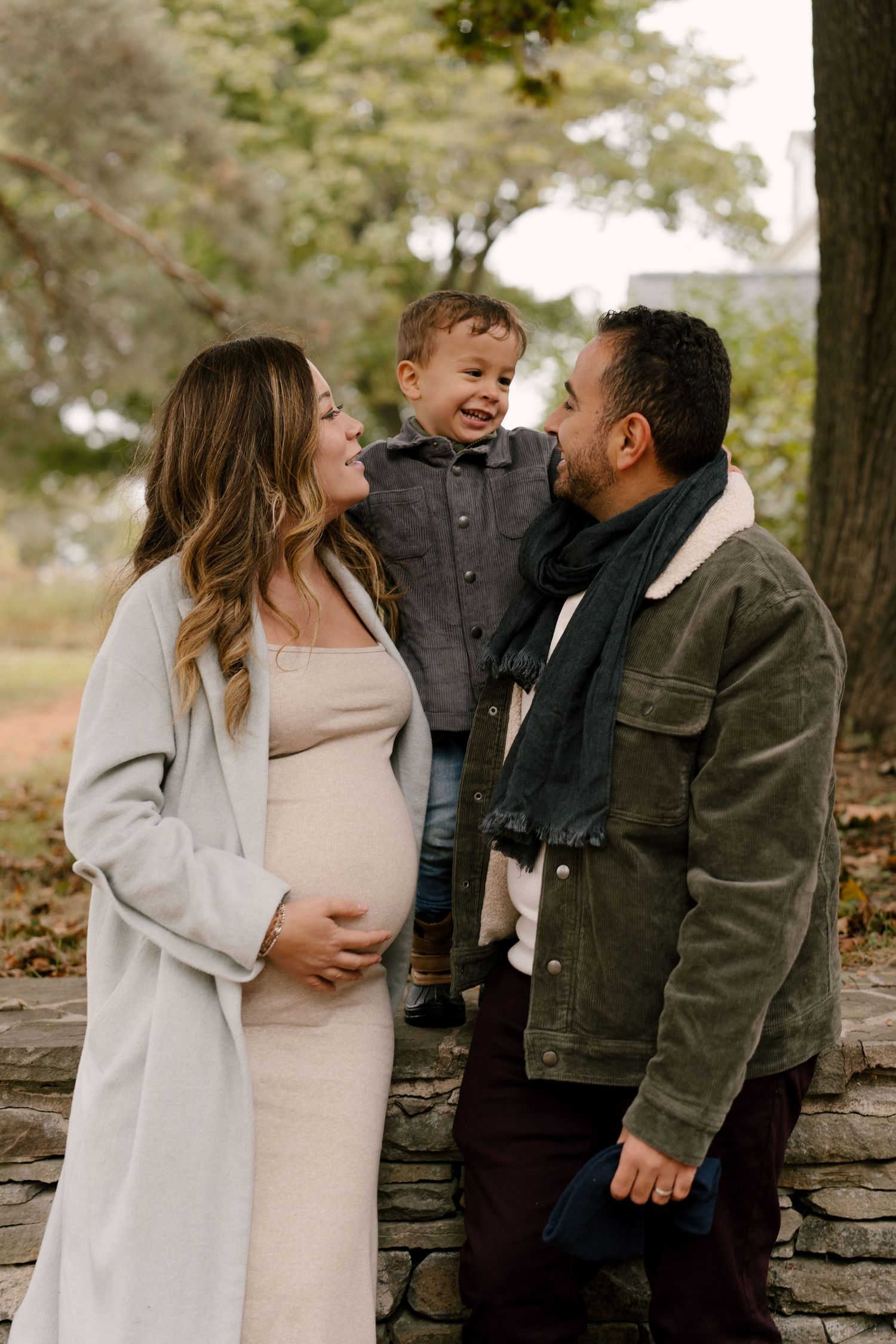 Pregnant mother and father smiling at laughing toddler between them near stone wall outdoors.