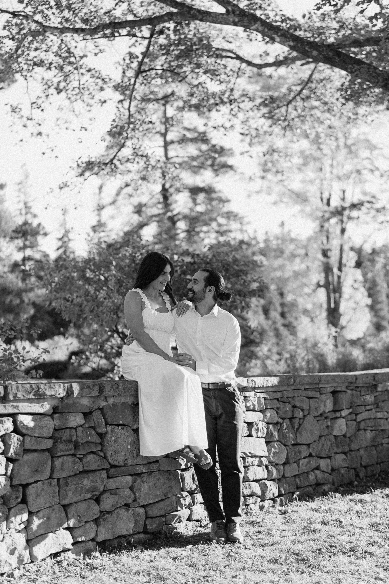 Black and white photo of couple sitting on a stone wall surrounded by trees outdoors at Scottsdale Farms.