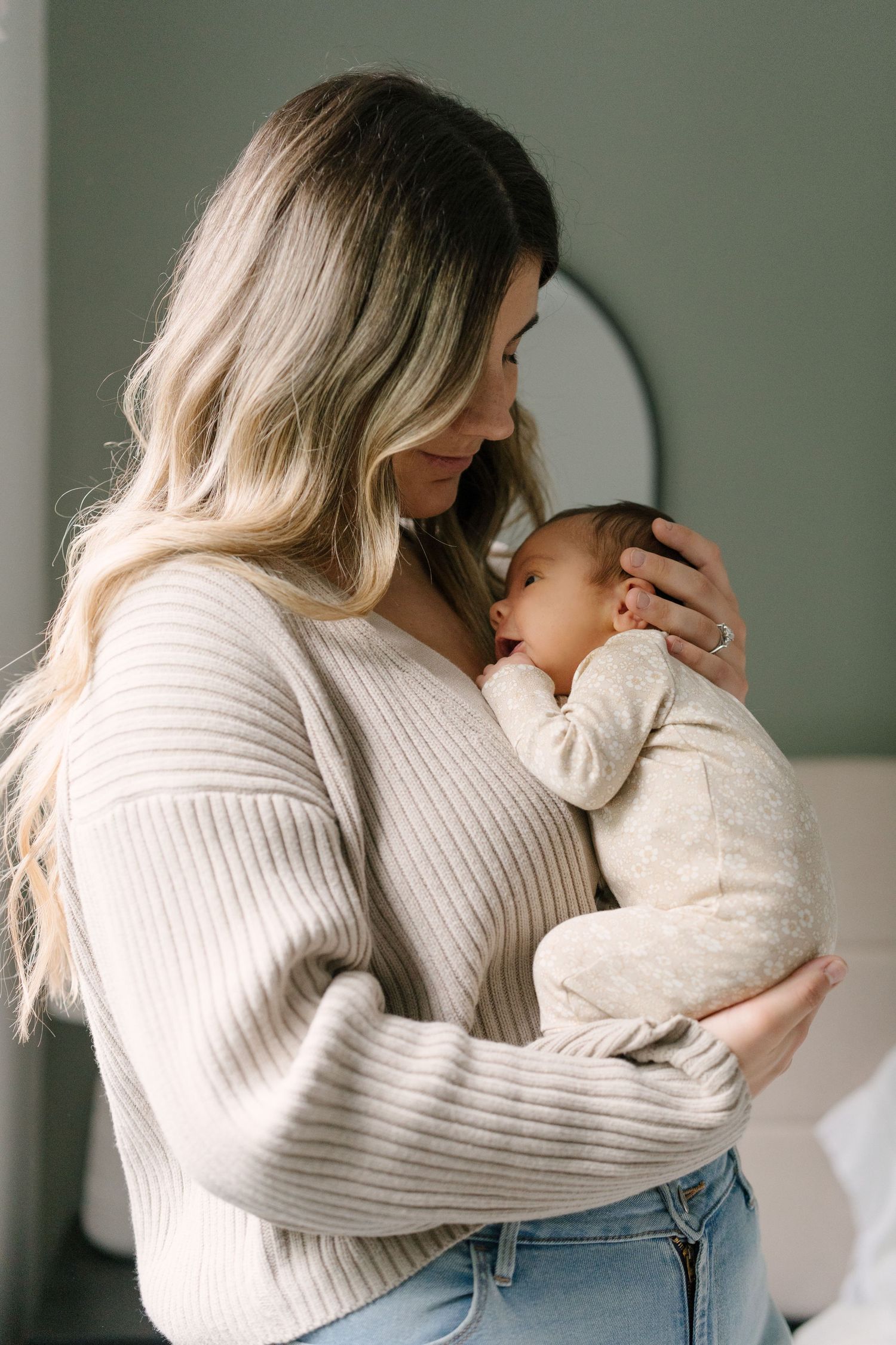 A woman in a cream ribbed sweater tenderly holds and gazes at her newborn baby in a softly lit room.