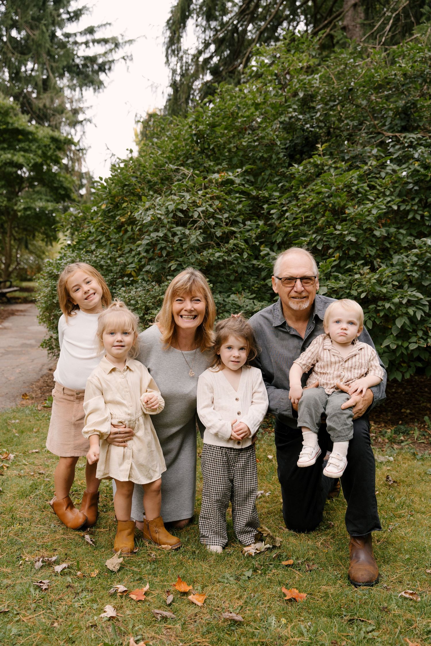 Grandparents posing with four young grandchildren in front of green garden shrubs.