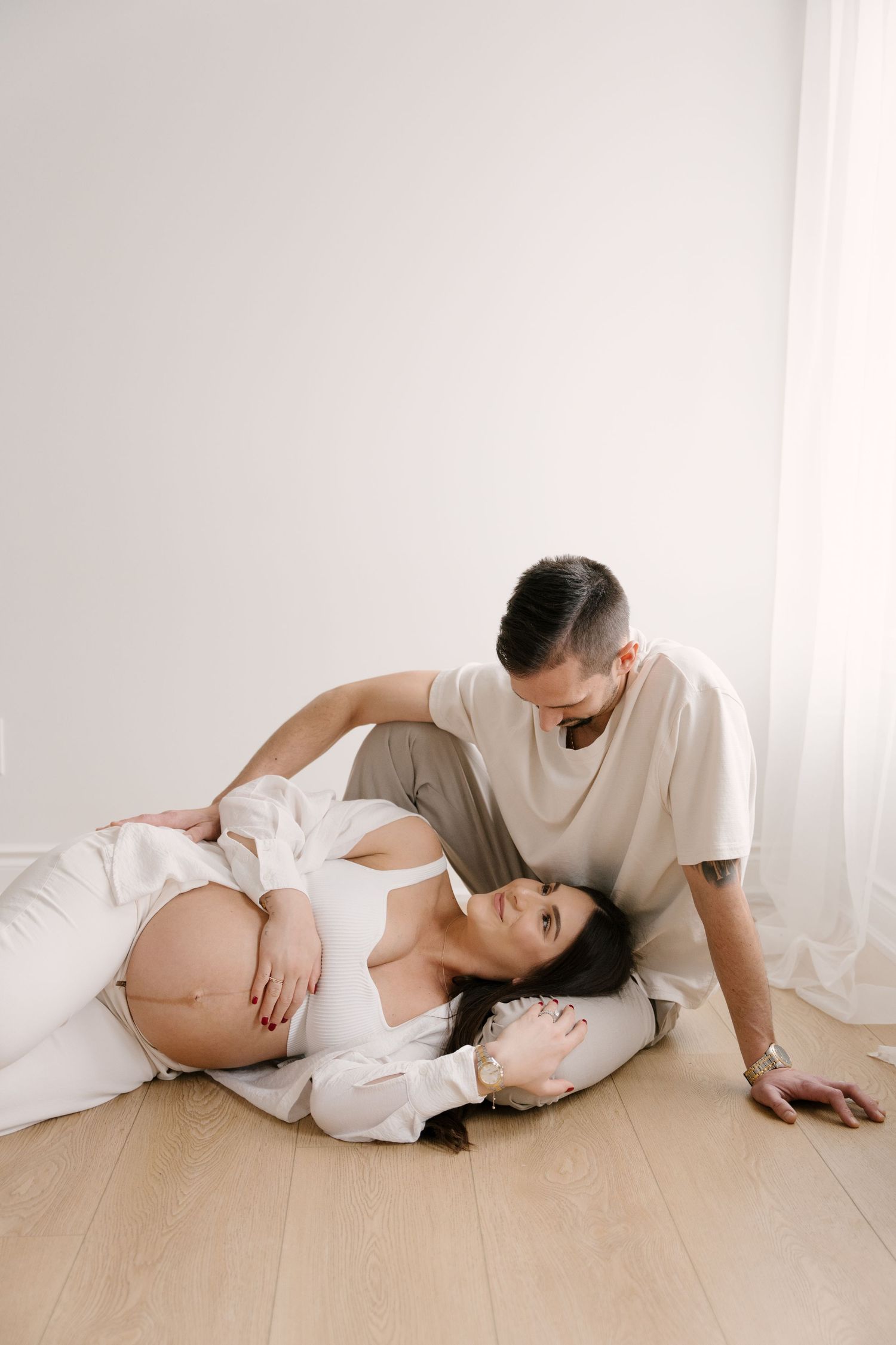 Pregnant woman lying on floor with head on mans lap during intimate maternity photo session in white room.