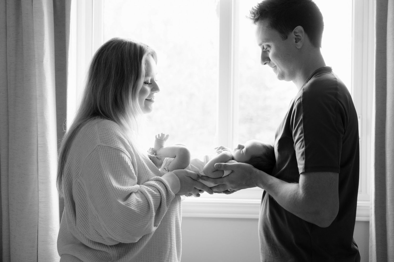 Black and white image of new parents facing each other by window passing their newborn baby.