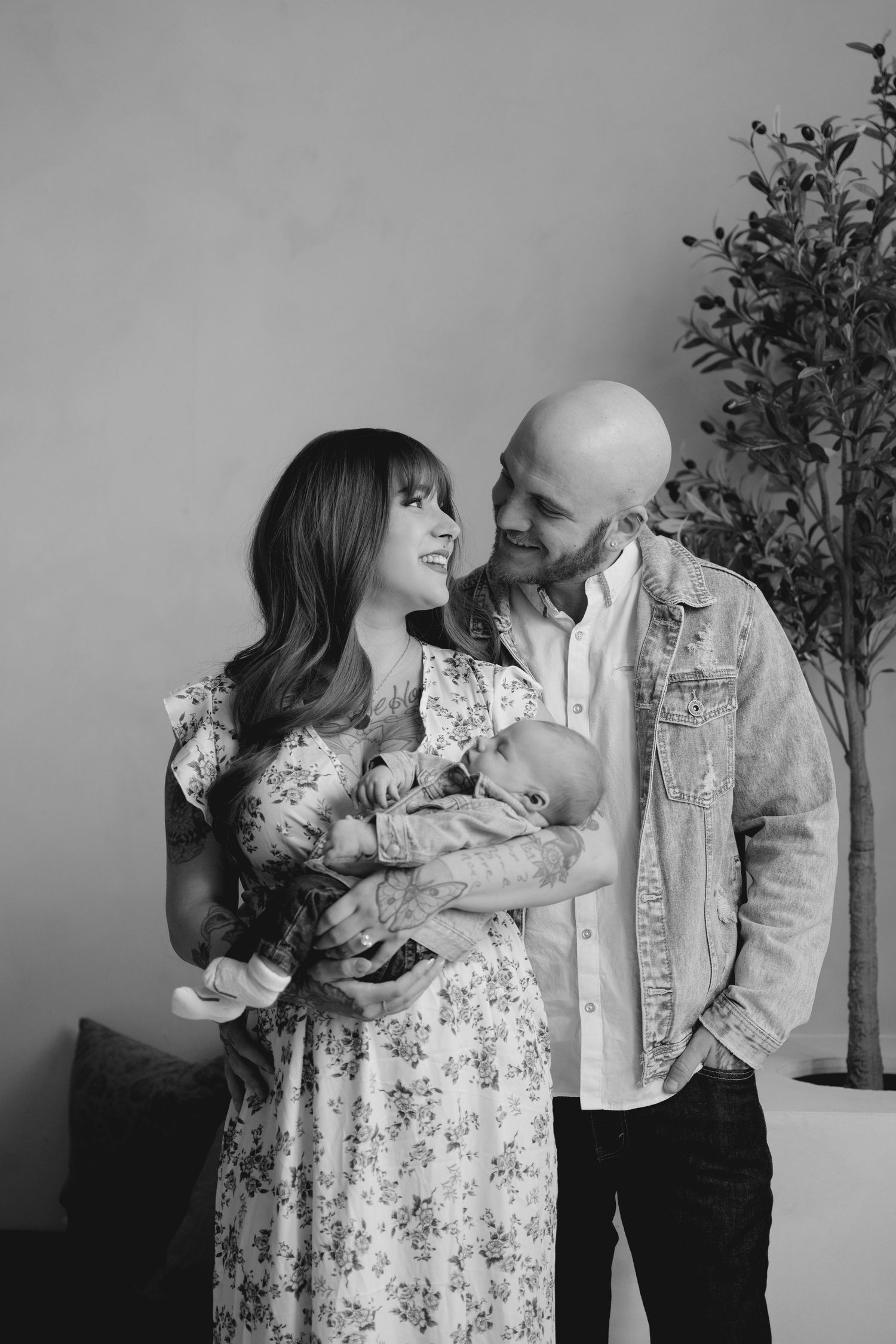 Black and white portrait of a couple in floral dress and denim jacket lovingly holding their newborn.