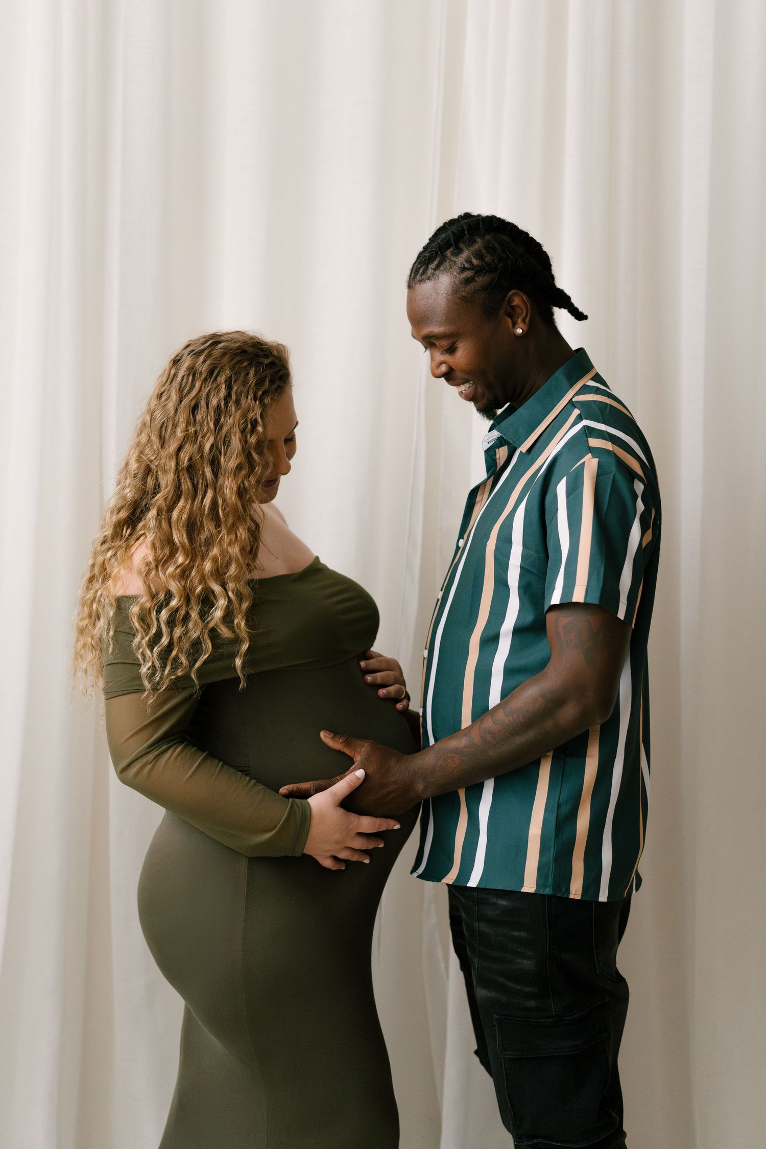 Interracial expecting couple smiling and touching pregnant belly against white curtain background.