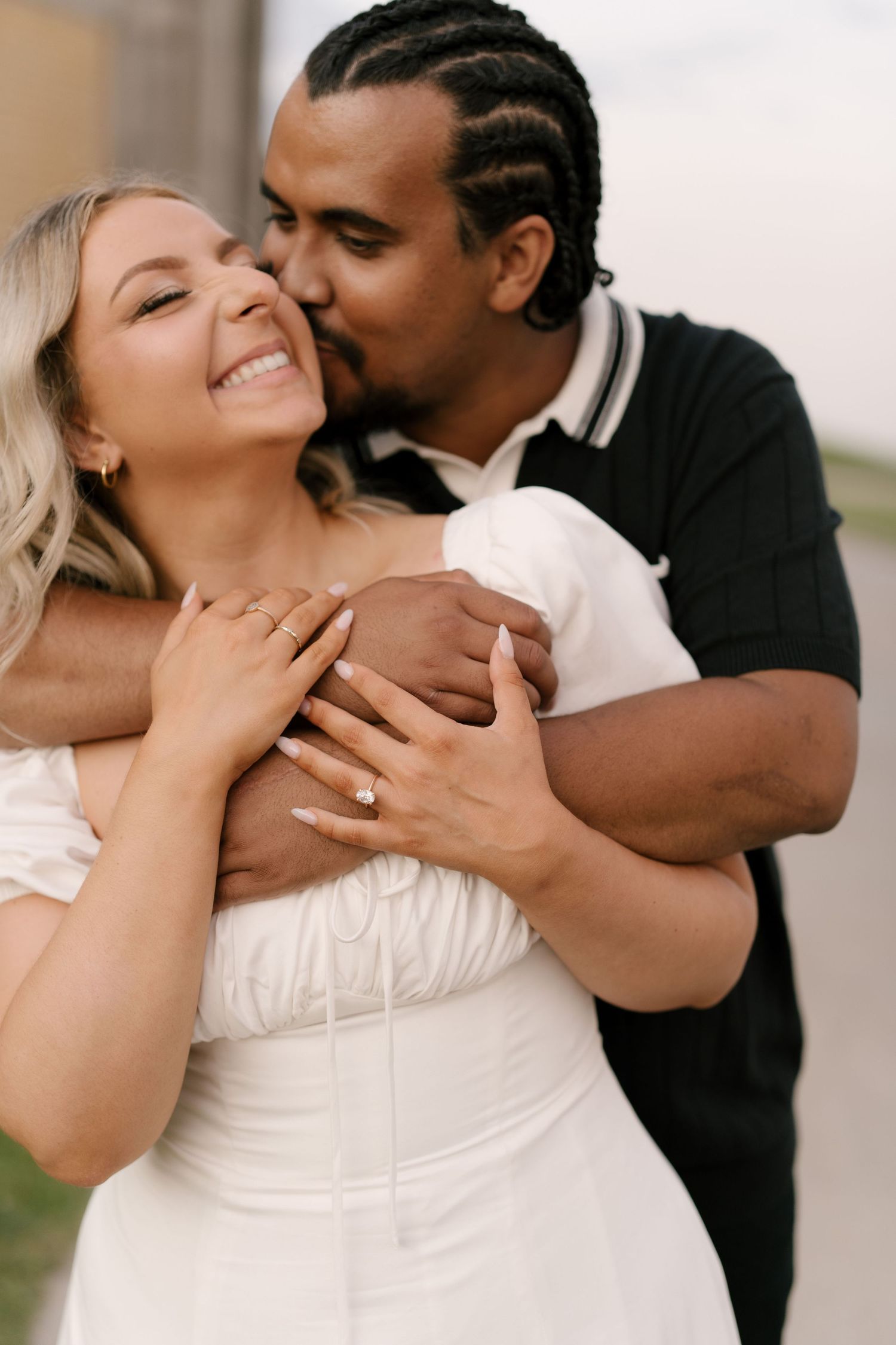 A couple shares a tender moment as the man kisses the womans cheek while hugging her from behind outdoors.