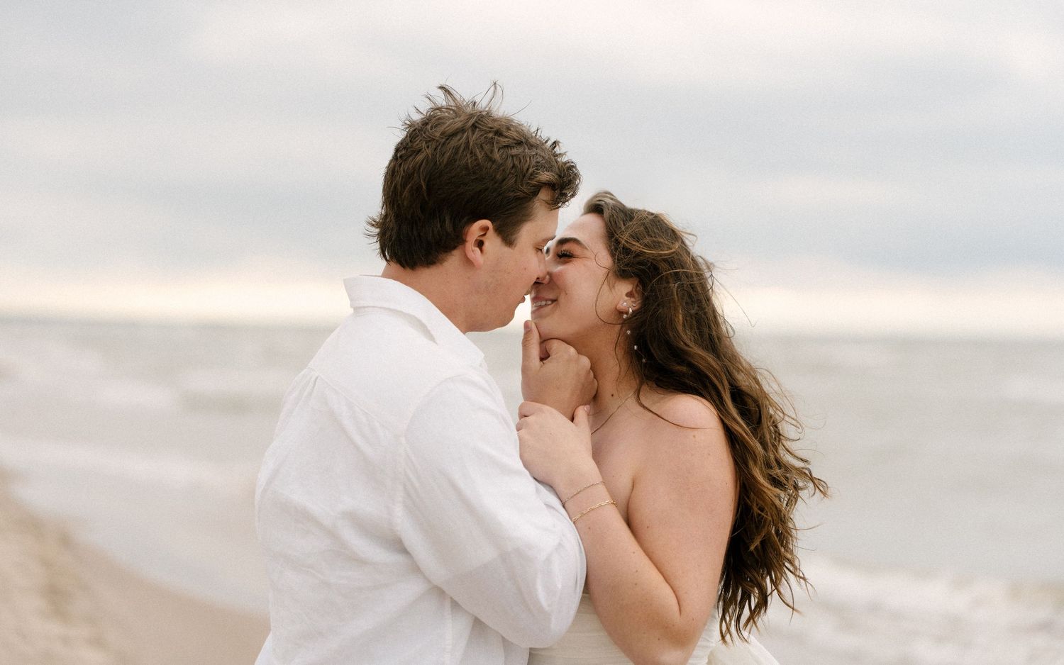 Couple sharing a close romantic moment nearly kissing on a windy beach with ocean backdrop.