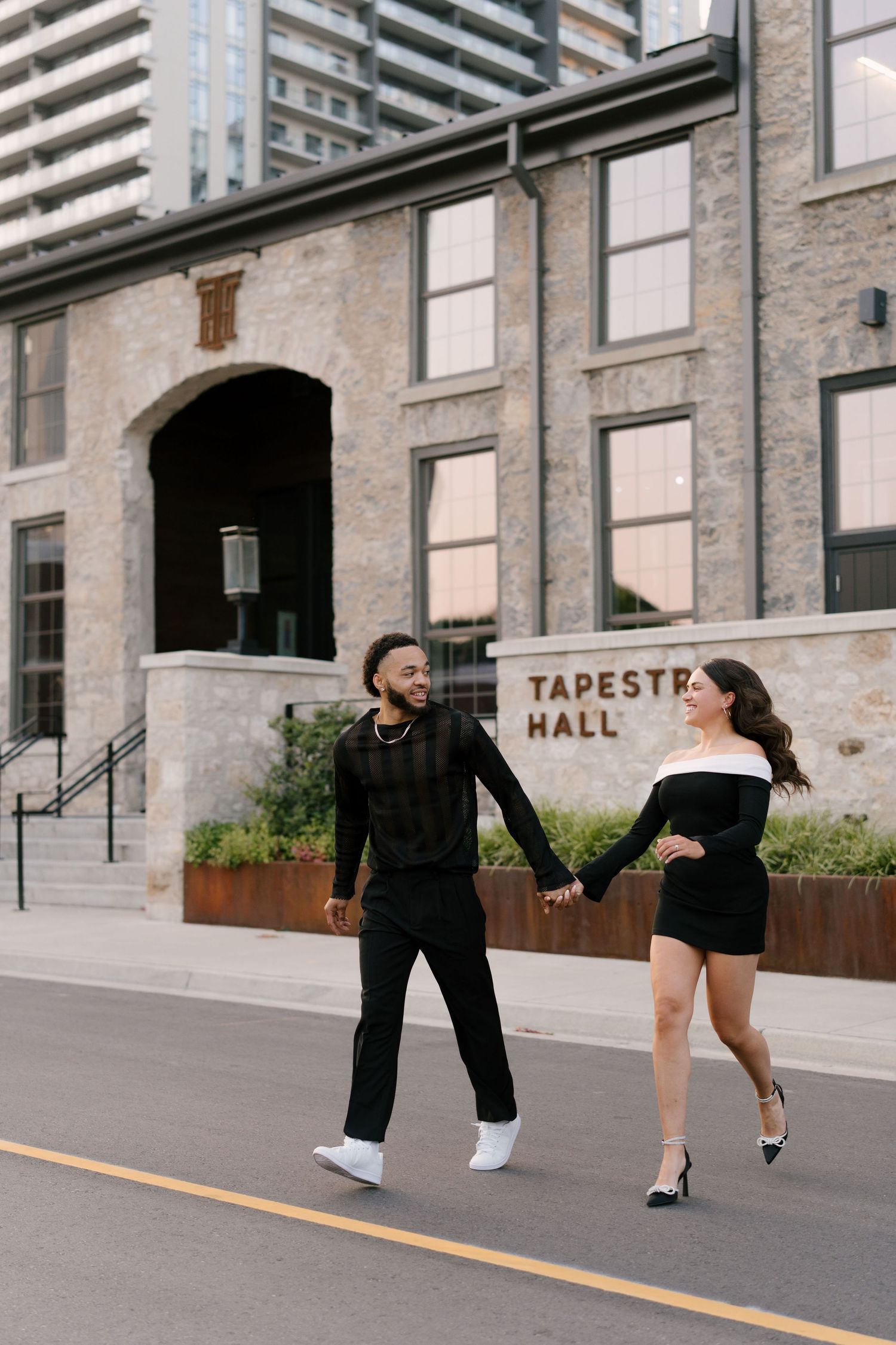 Stylish couple in black outfits hold hands walking in front of Tapestry Hall building.