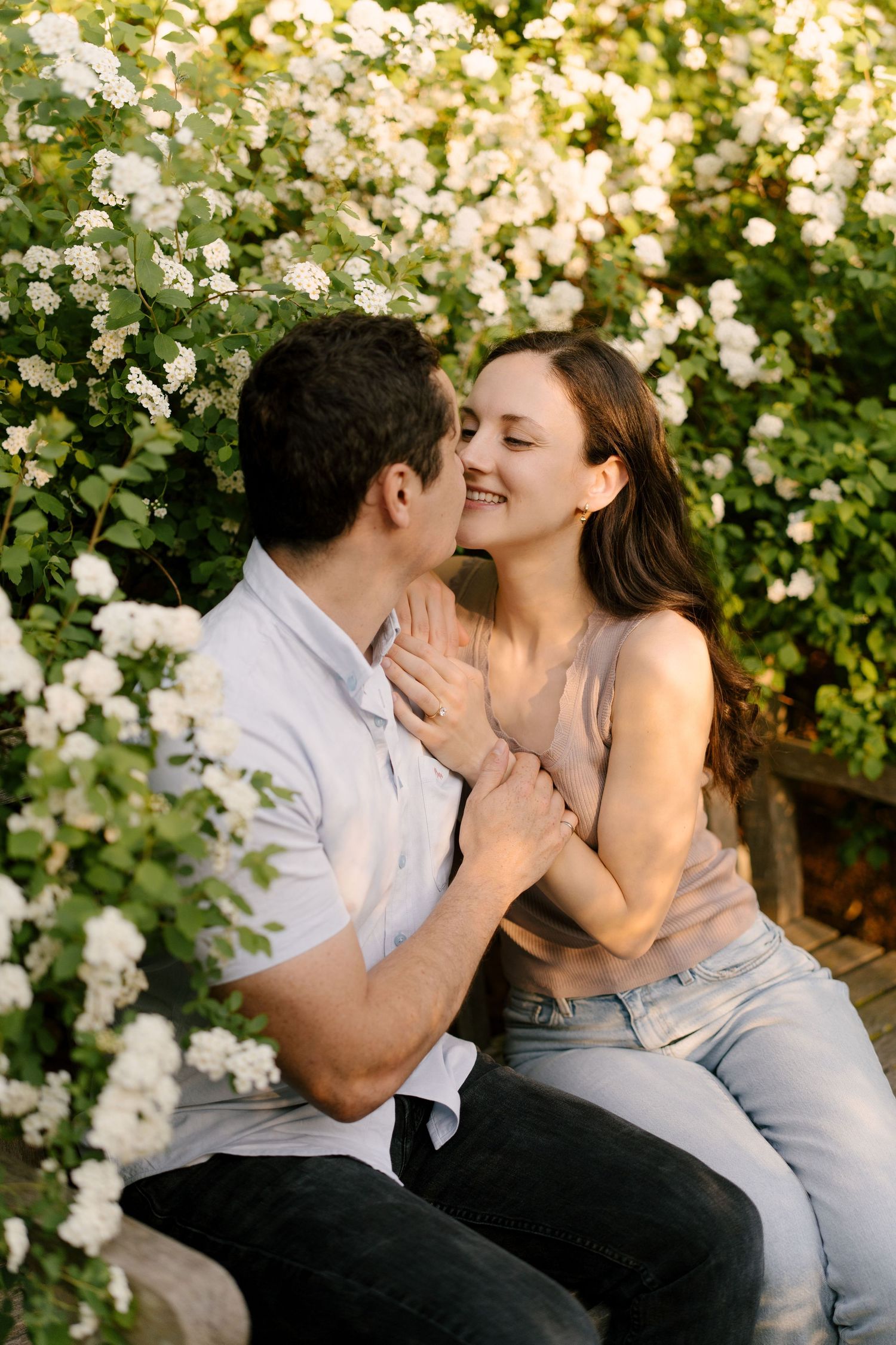 Romantic couple sitting on a bench surrounded by white flowering bushes, sharing a tender moment at sunset.