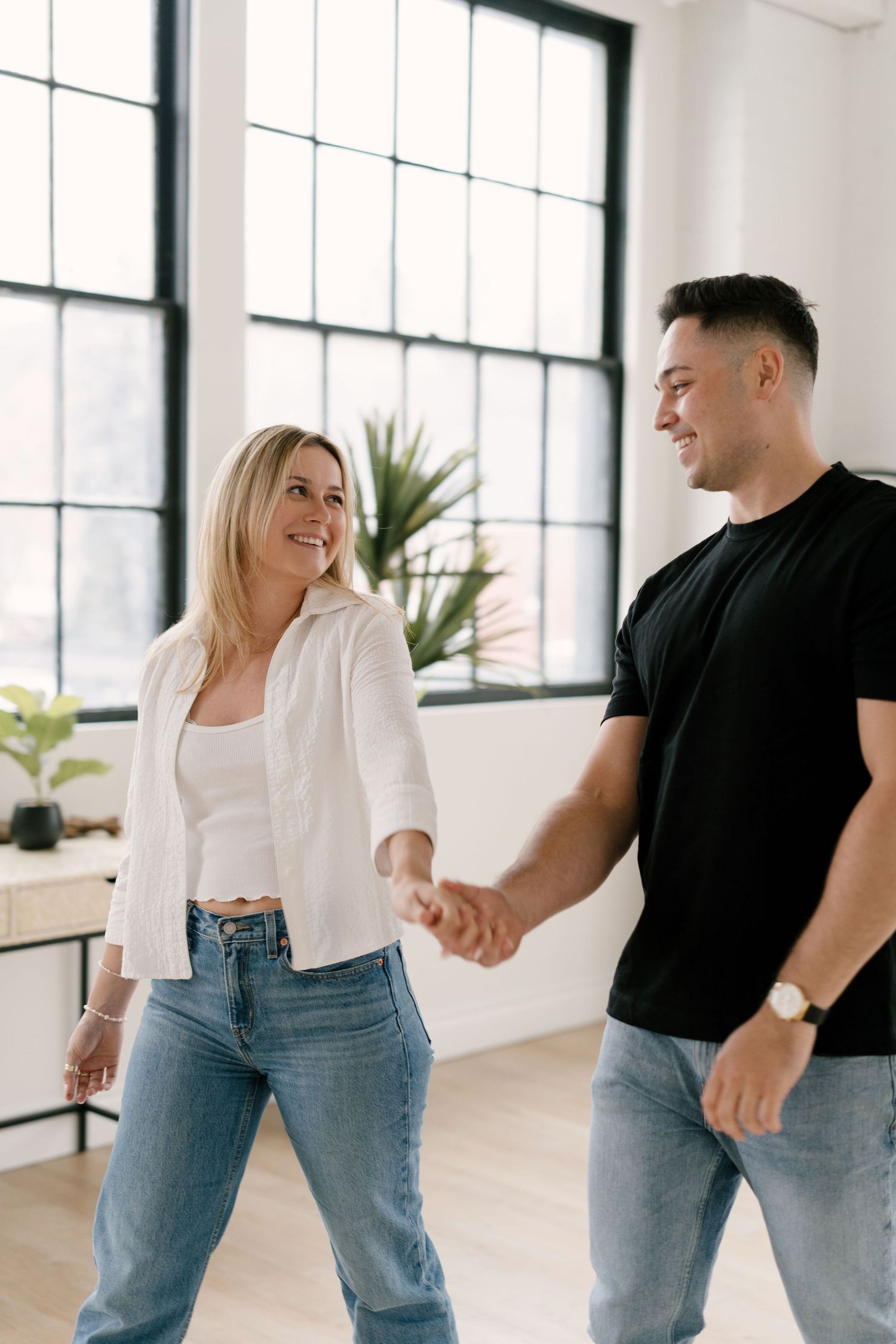 Smiling couple holding hands in a bright modern studio with large windows.