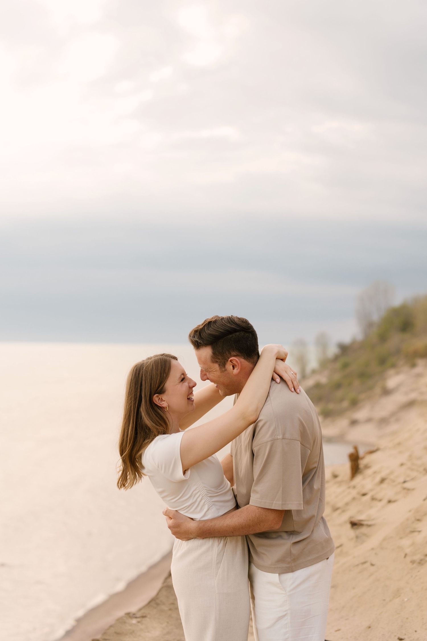 Couple embracing and smiling at each other on a sandy beach with cloudy sky.