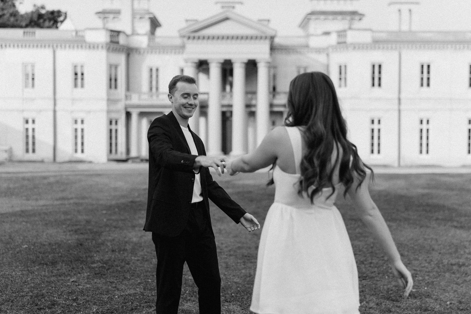 Black and white photo of couple holding hands dancing outside elegant mansion.