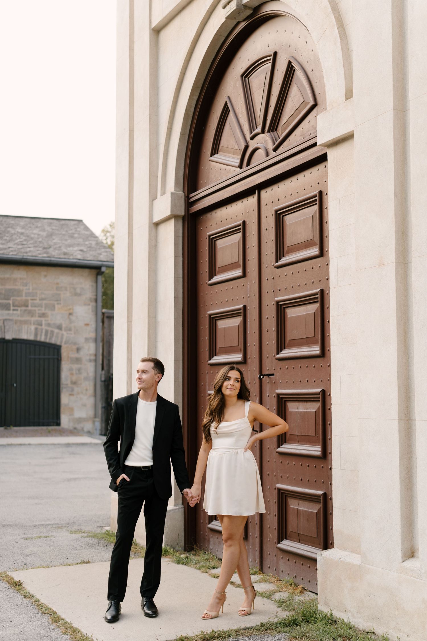 Couple holding hands in front of ornate wooden church doors, man in black suit, woman in white mini dress.