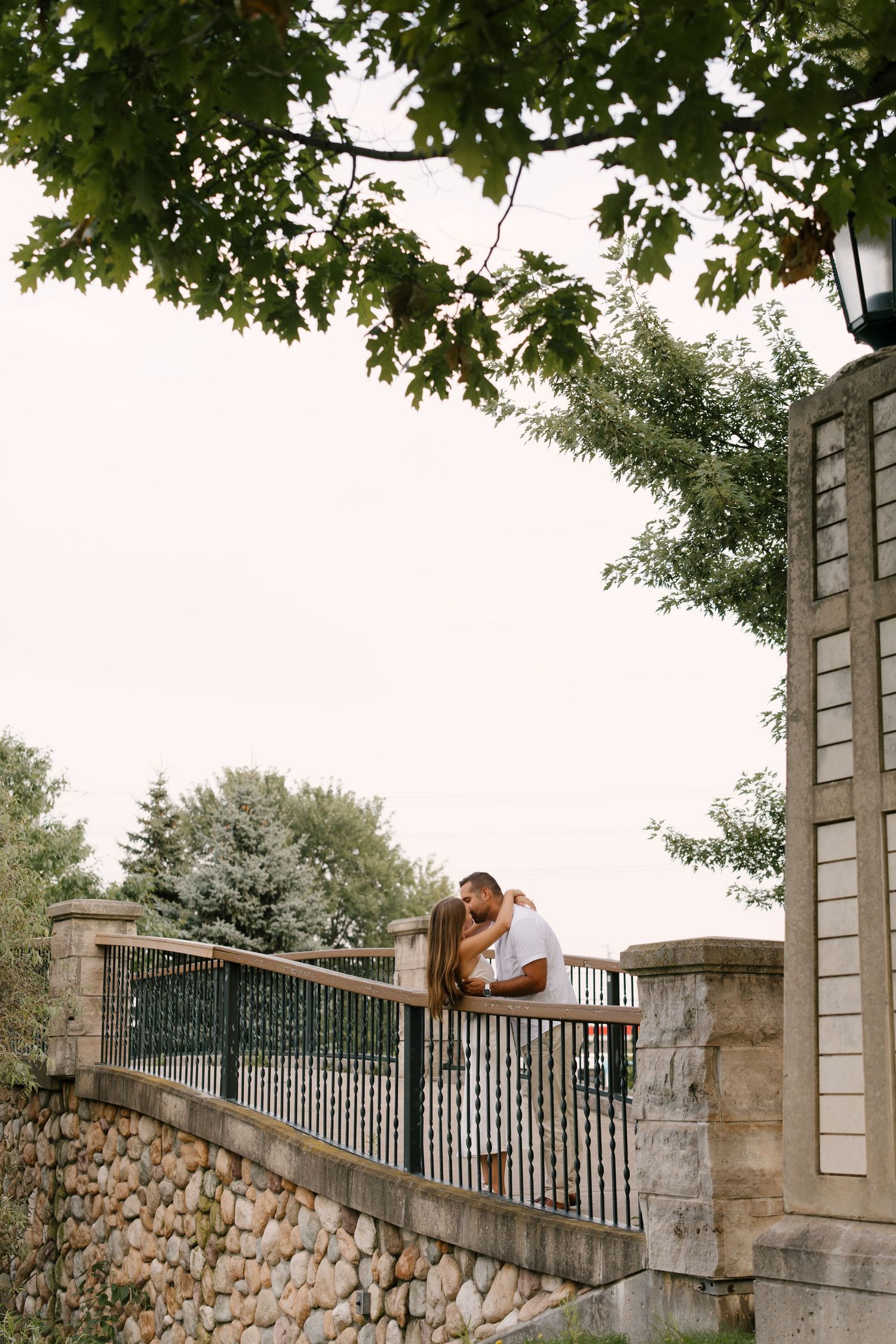 Romantic couple embracing on stone bridge balcony surrounded by green trees in soft natural light.