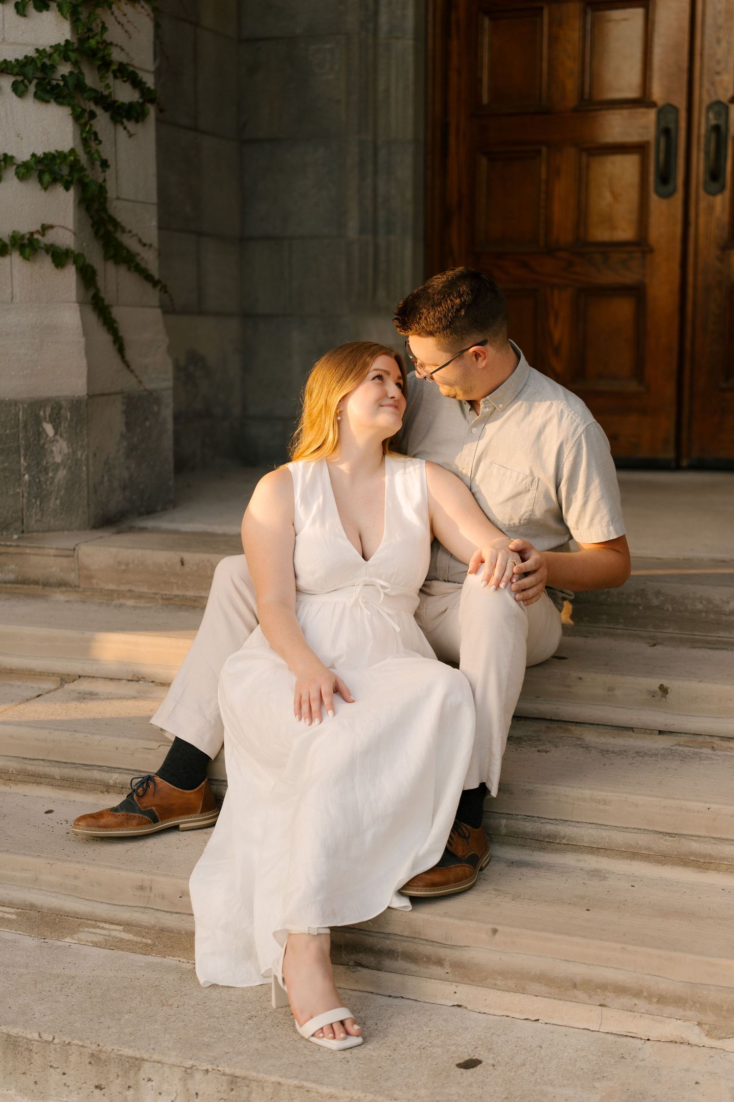 Couple sitting on stone steps, woman in white dress gazing up at man in light shirt during golden hour.