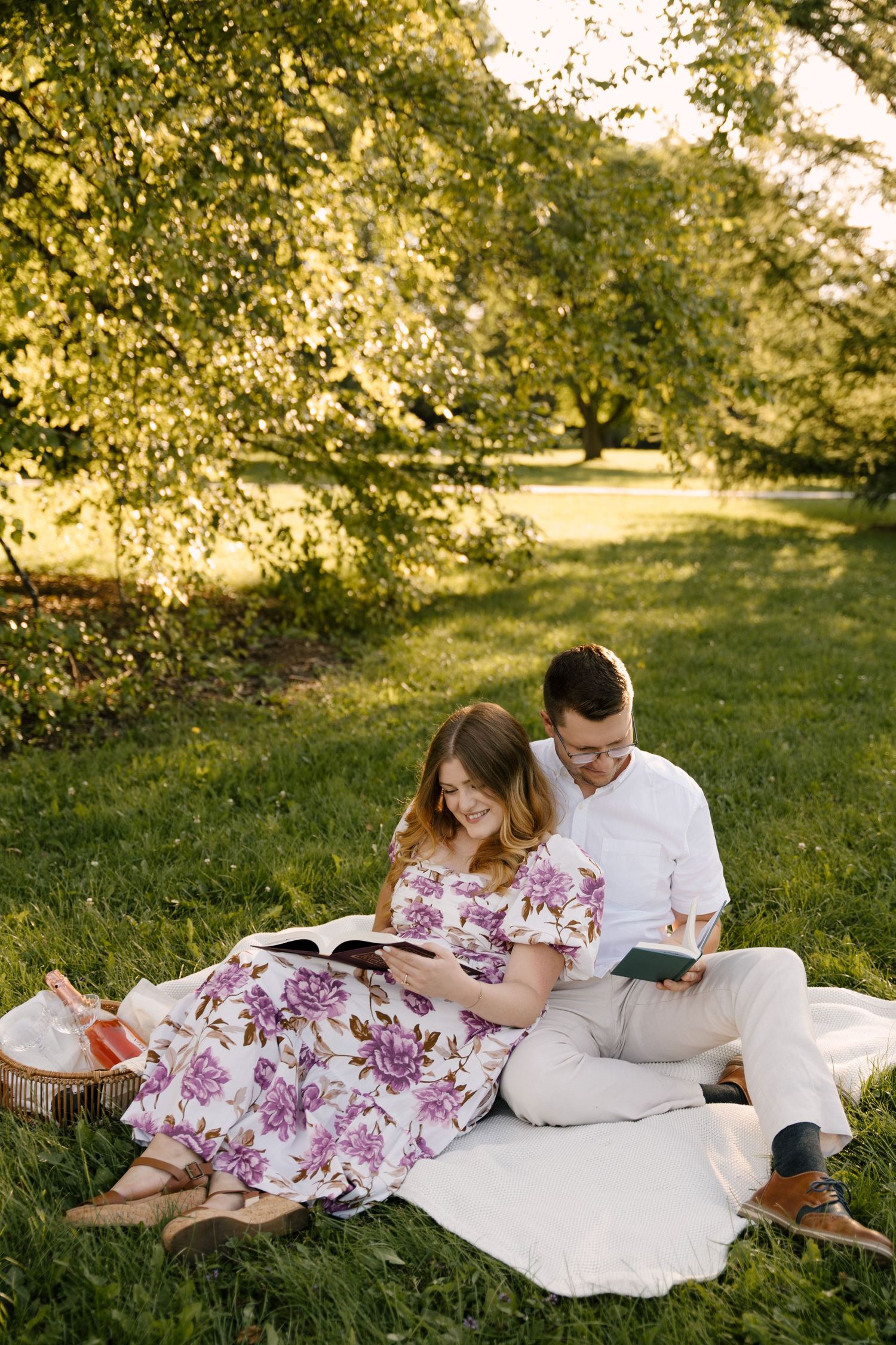 Couple reading books together on a white blanket under lush green trees in golden sunlight.