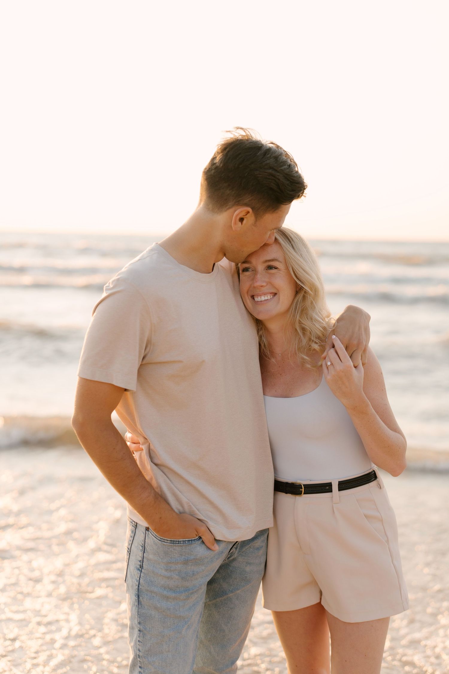 Couple embracing on sunlit beach at sunset, man kissing womans temple as she smiles.