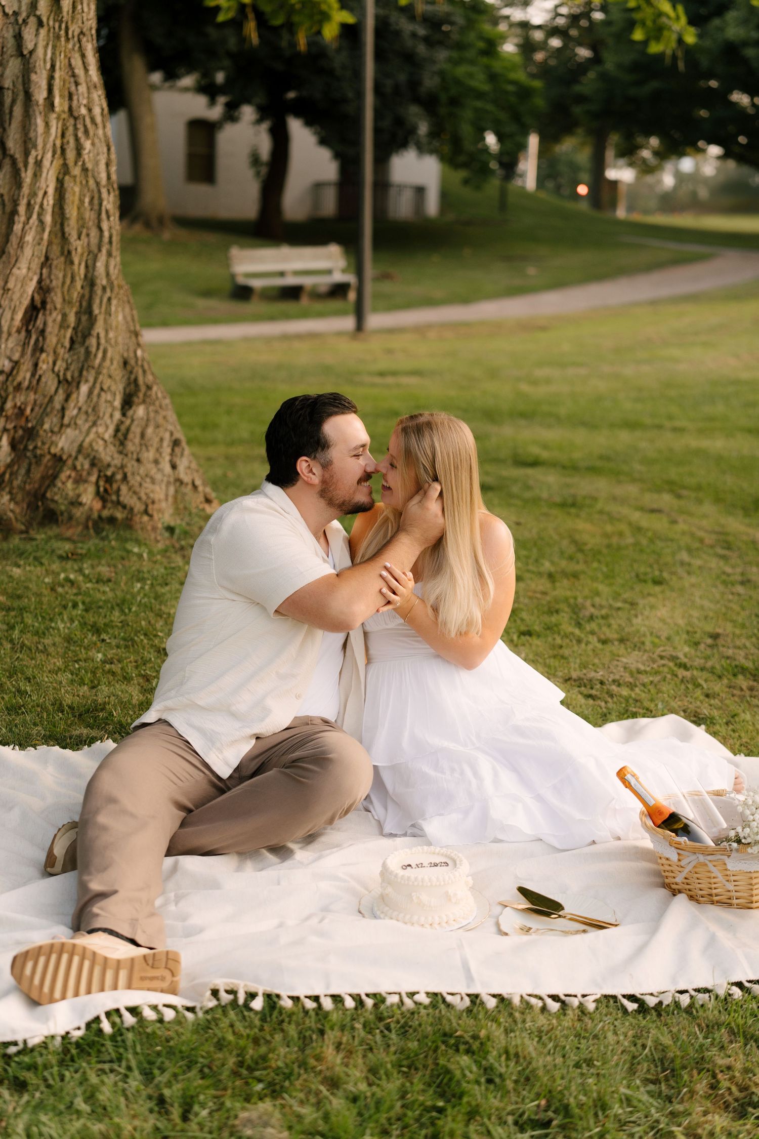 Couple sharing a romantic kiss on a white blanket during a picnic in a lush green park.