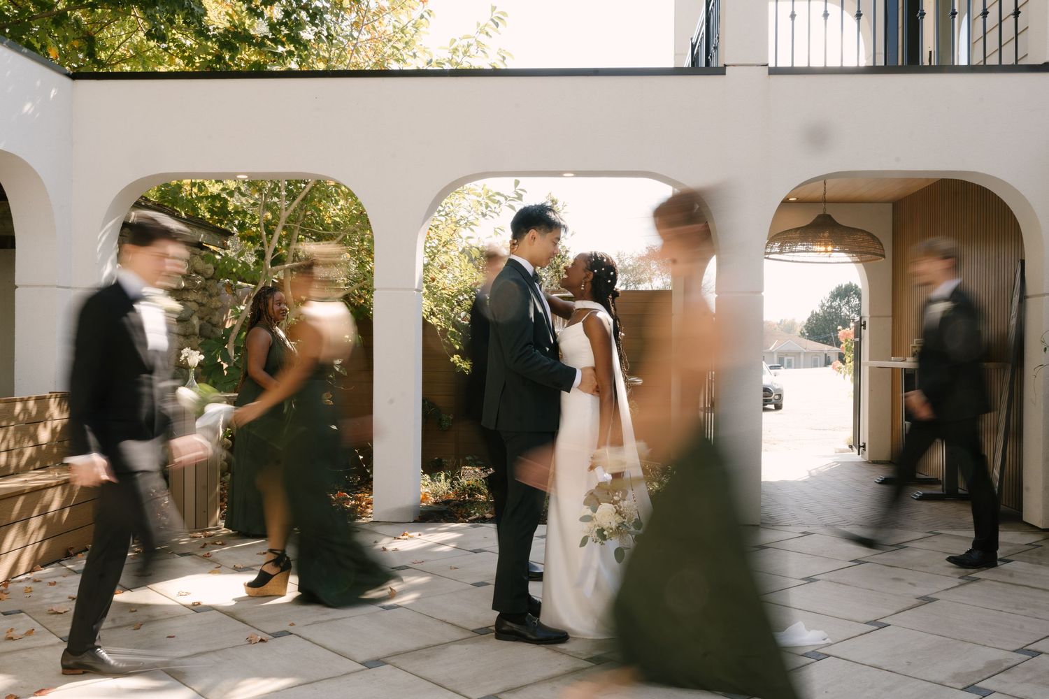 Wedding couple kisses under a white arch while the bridal party moves around them.