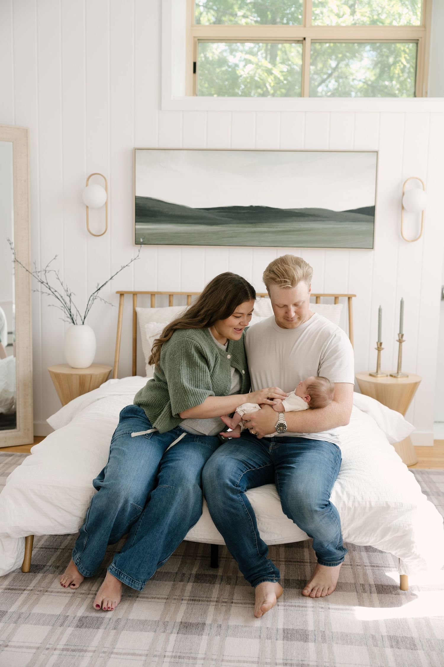 New parents sitting on bed smiling at newborn baby in bright bedroom.