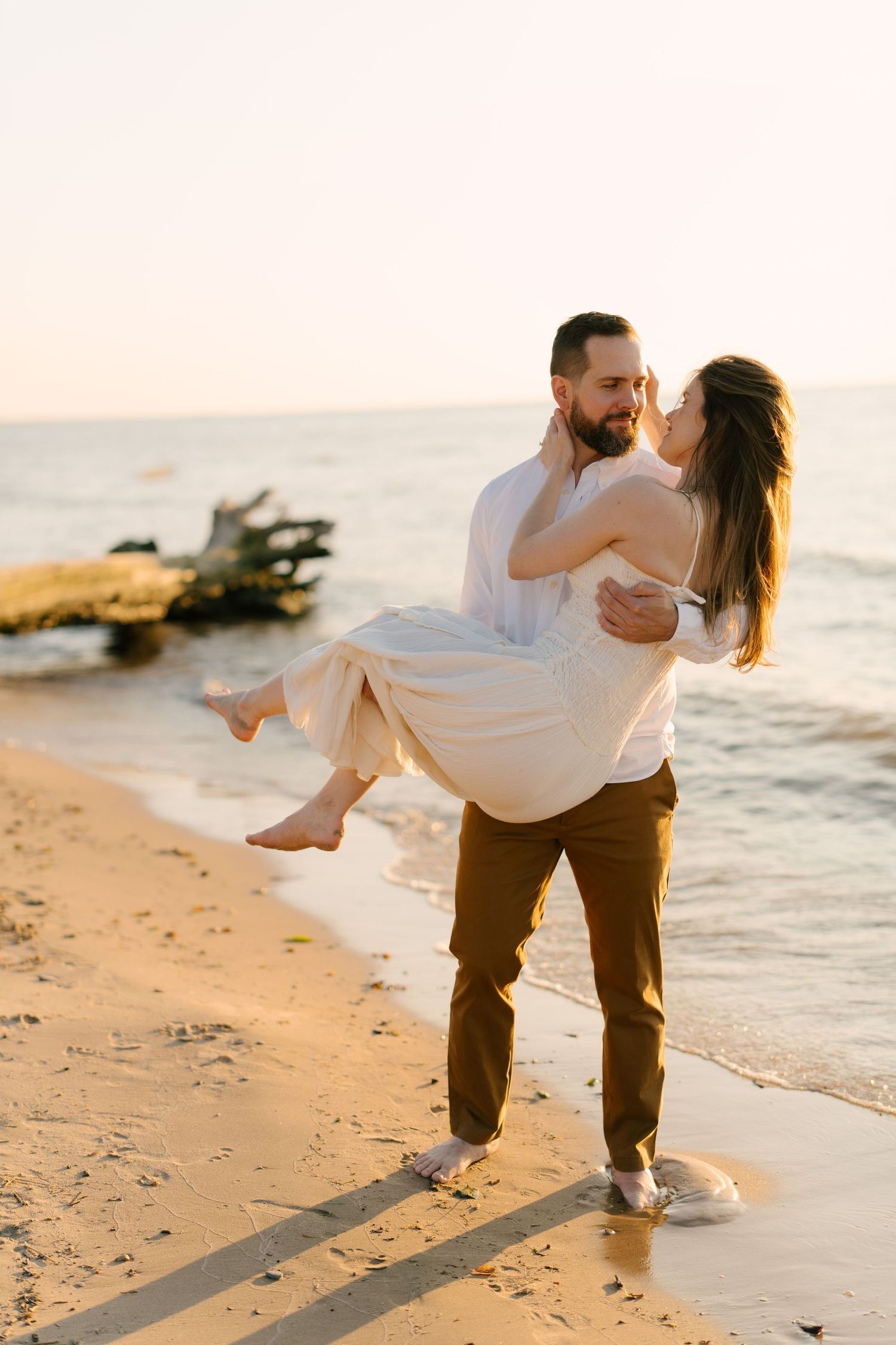 Man carrying woman in white dress along a golden sandy beach at sunset with ocean waves in the background.