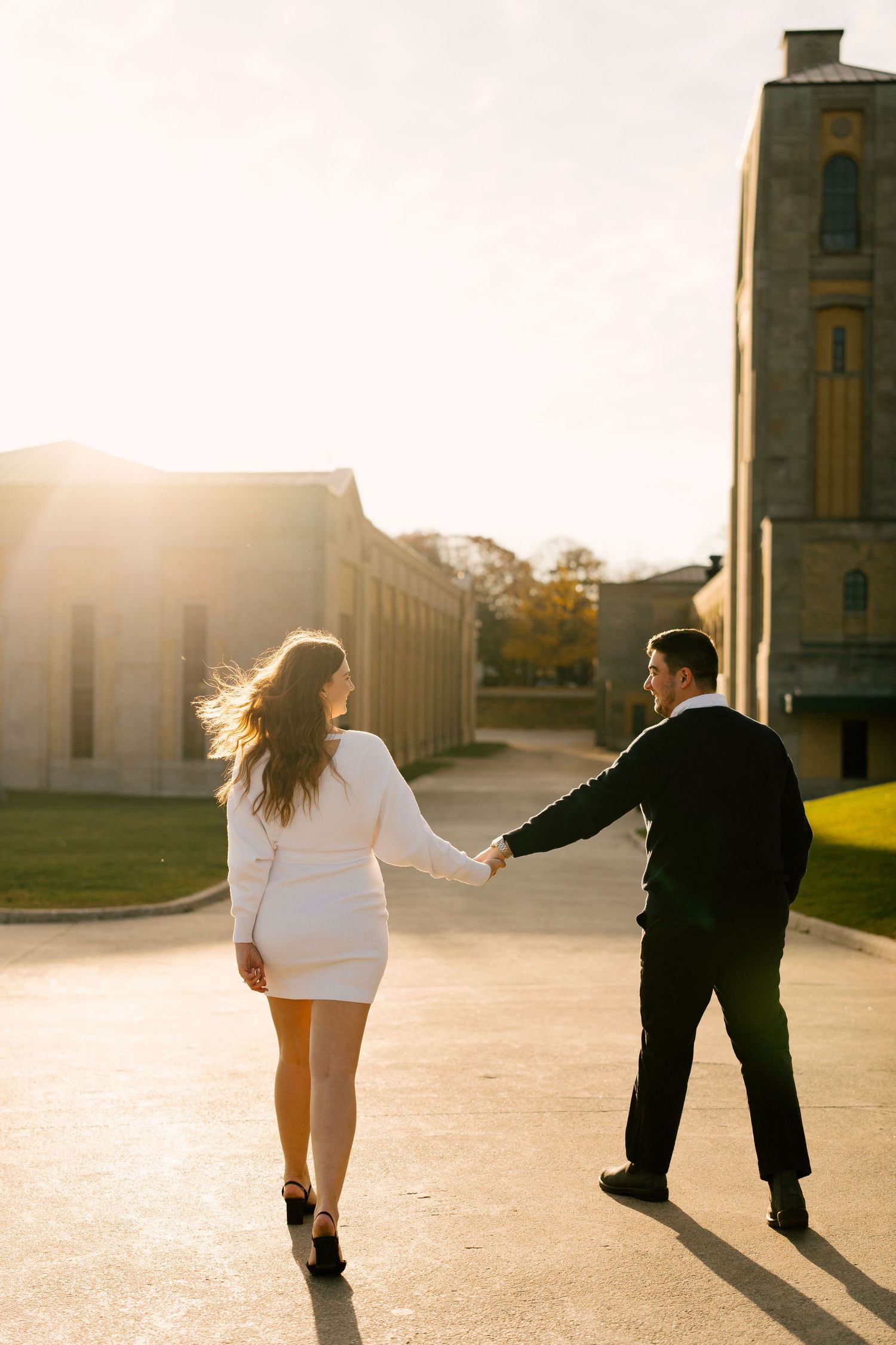 Couple holding hands walking toward golden sunset between historic stone buildings.