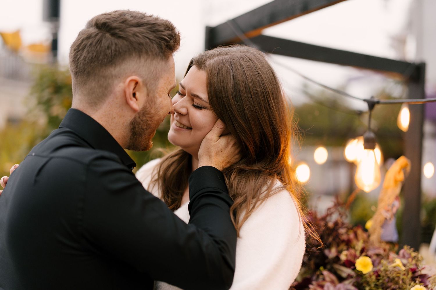 Romantic couple about to kiss on a rooftop with warm string lights glowing behind them.