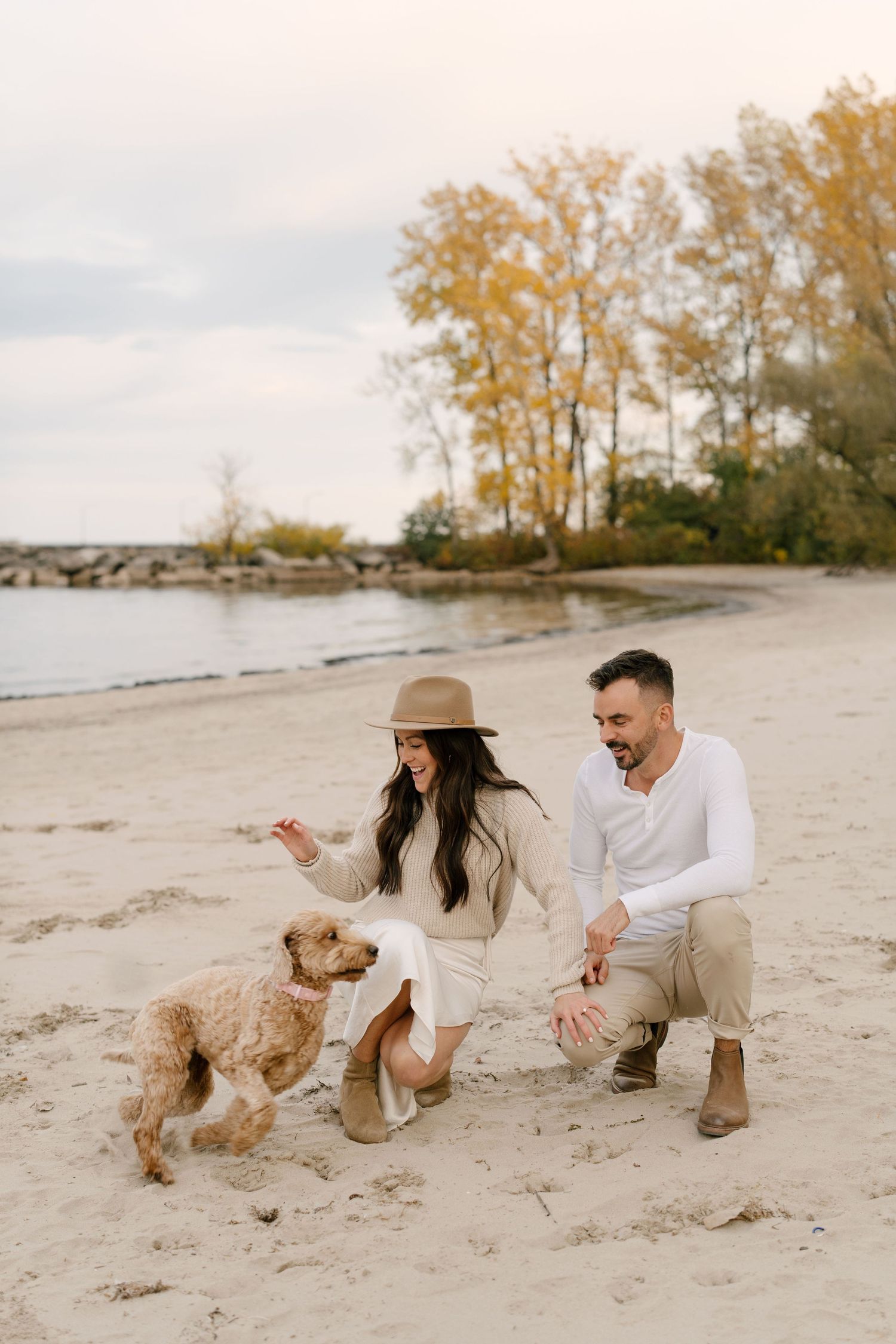 Couple playing with their fluffy dog on a sandy beach during autumn, with colorful trees and calm water in the background.