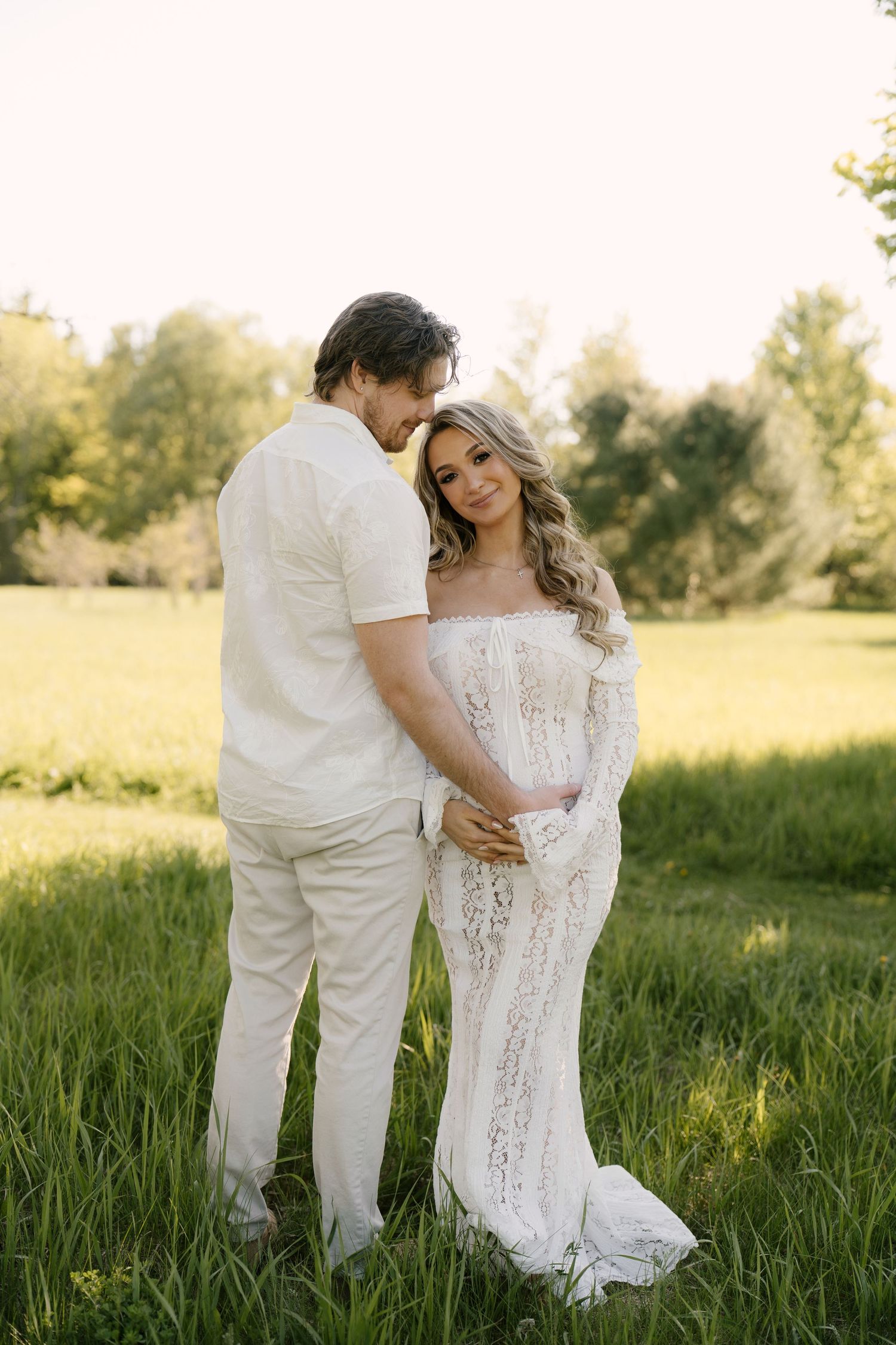 Couple embracing in golden sunlight on green grass, pregnant woman in elegant white lace maternity dress.